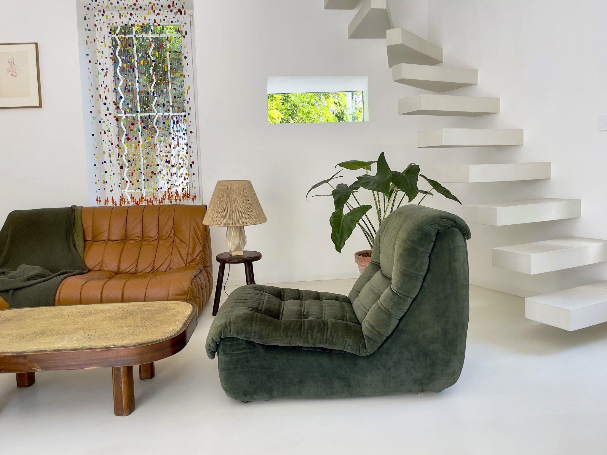 A modern minimalist living room features a brown leather sofa, a plush green armchair, a wooden coffee table, and a side table with a lamp. A large plant is nearby, and a floating staircase leads to the upper floor. Artwork and a colorful curtain adorn the wall.