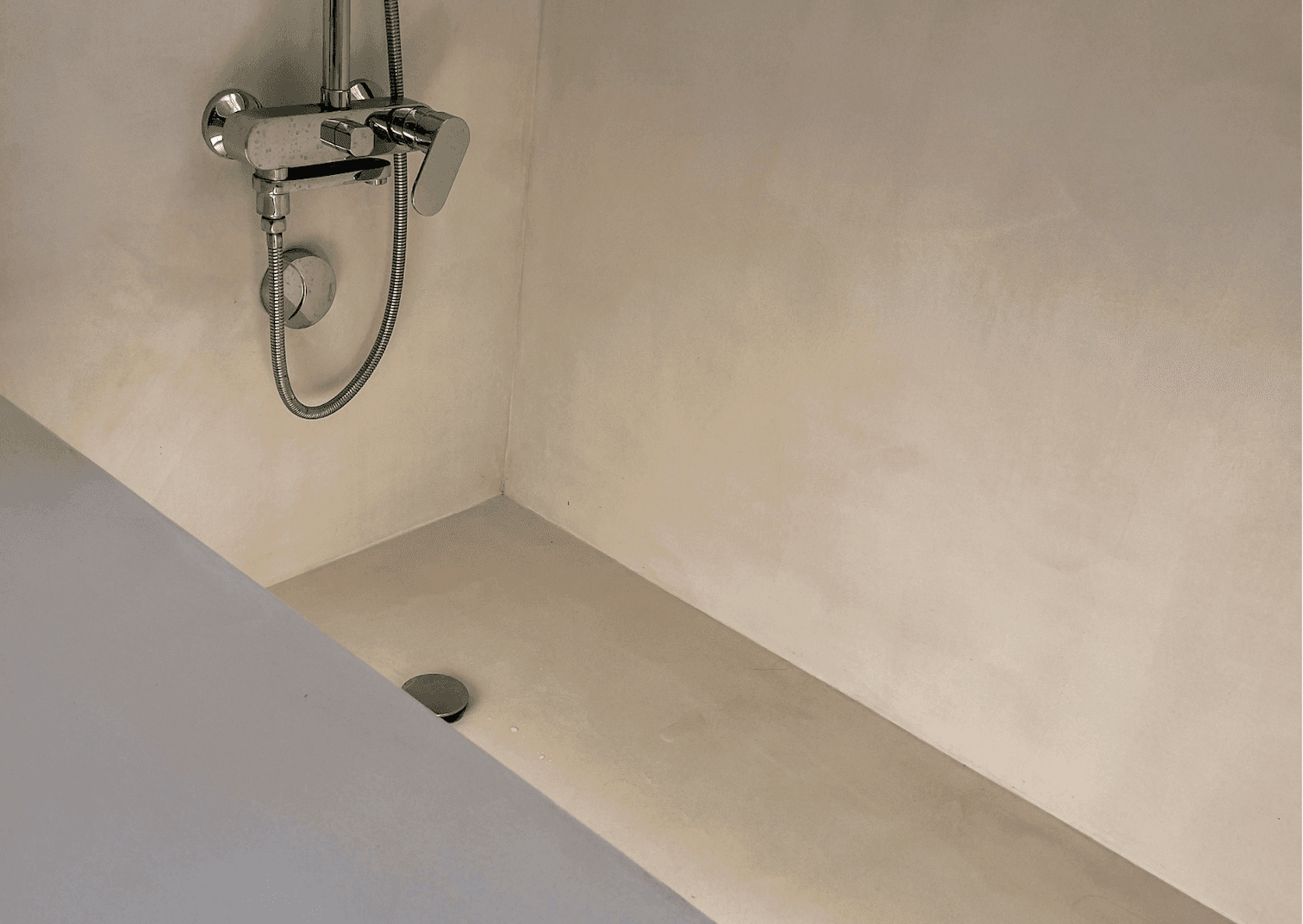 A minimalist shower features chrome wall-mounted fixtures, including a showerhead and a flexible hose. The shower has smooth cream-colored walls and floor, with a subtle drain located on the floor near the corner.