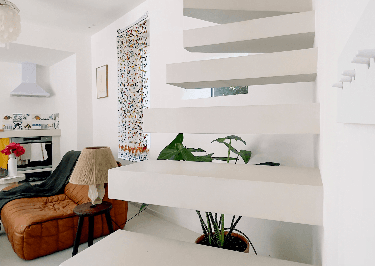 This is a modern, minimalist living room featuring floating white stairs, a brown leather pouf with a cozy throw, a floor lamp with a beige shade, and a large potted plant. The kitchen corner with decorative tiles on the backsplash is visible in the background.