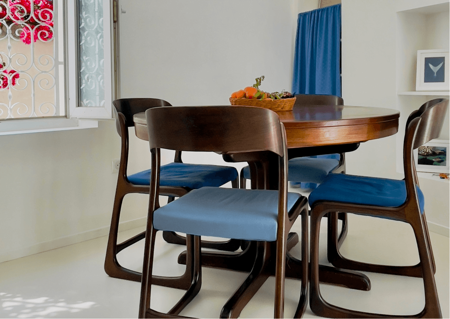 A round wooden dining table topped with a fruit basket is surrounded by four wooden chairs with blue cushions. The room features a white wall with a small window framed by ornate white grilles and blue curtains, along with a shelf in the background.