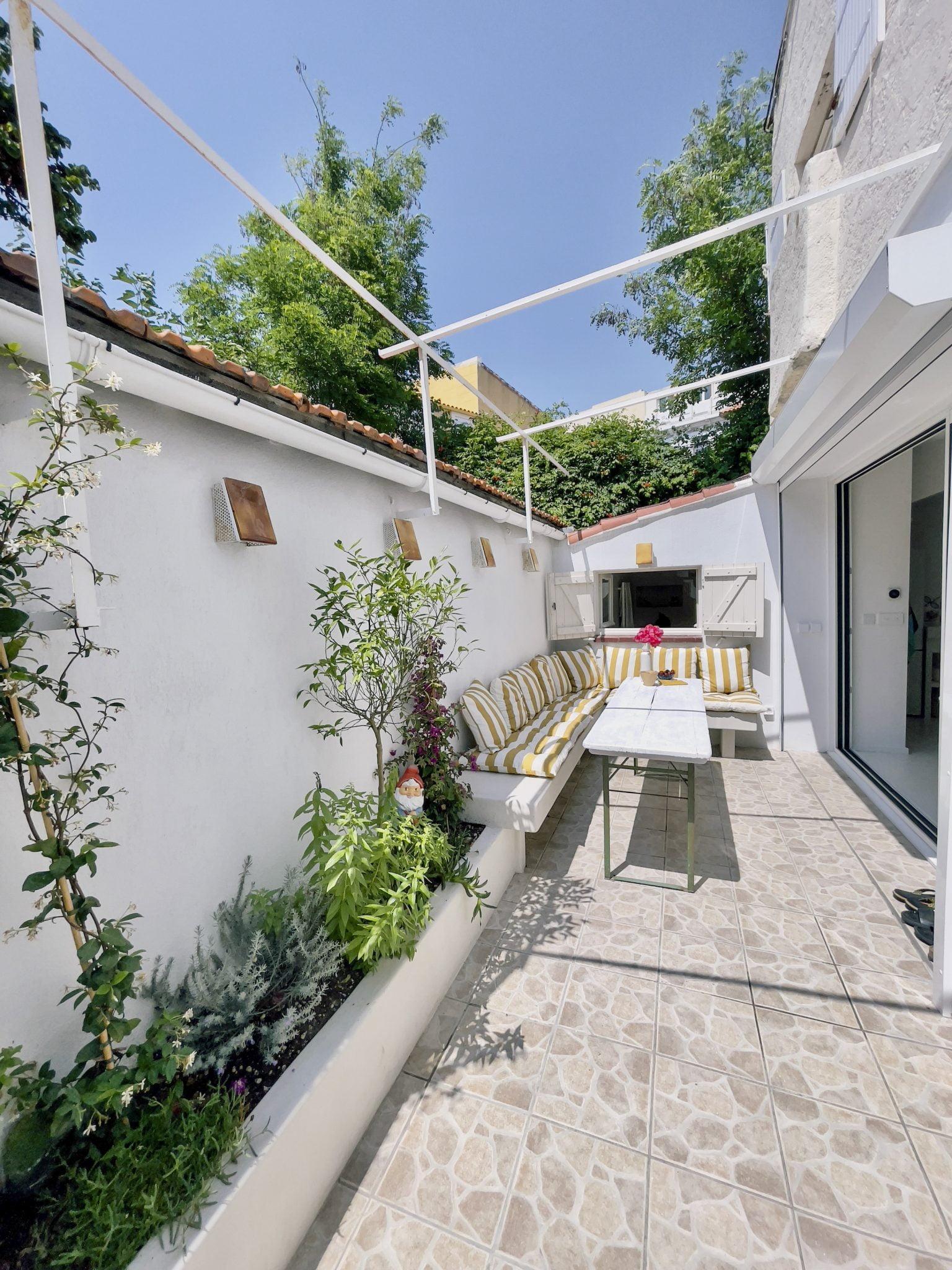 A bright outdoor patio featuring a cushioned seating area with yellow and white striped cushions along one wall. The space has a white stone-tiled floor, green plants in raised beds, a rectangular white table, and an open sky visible above.