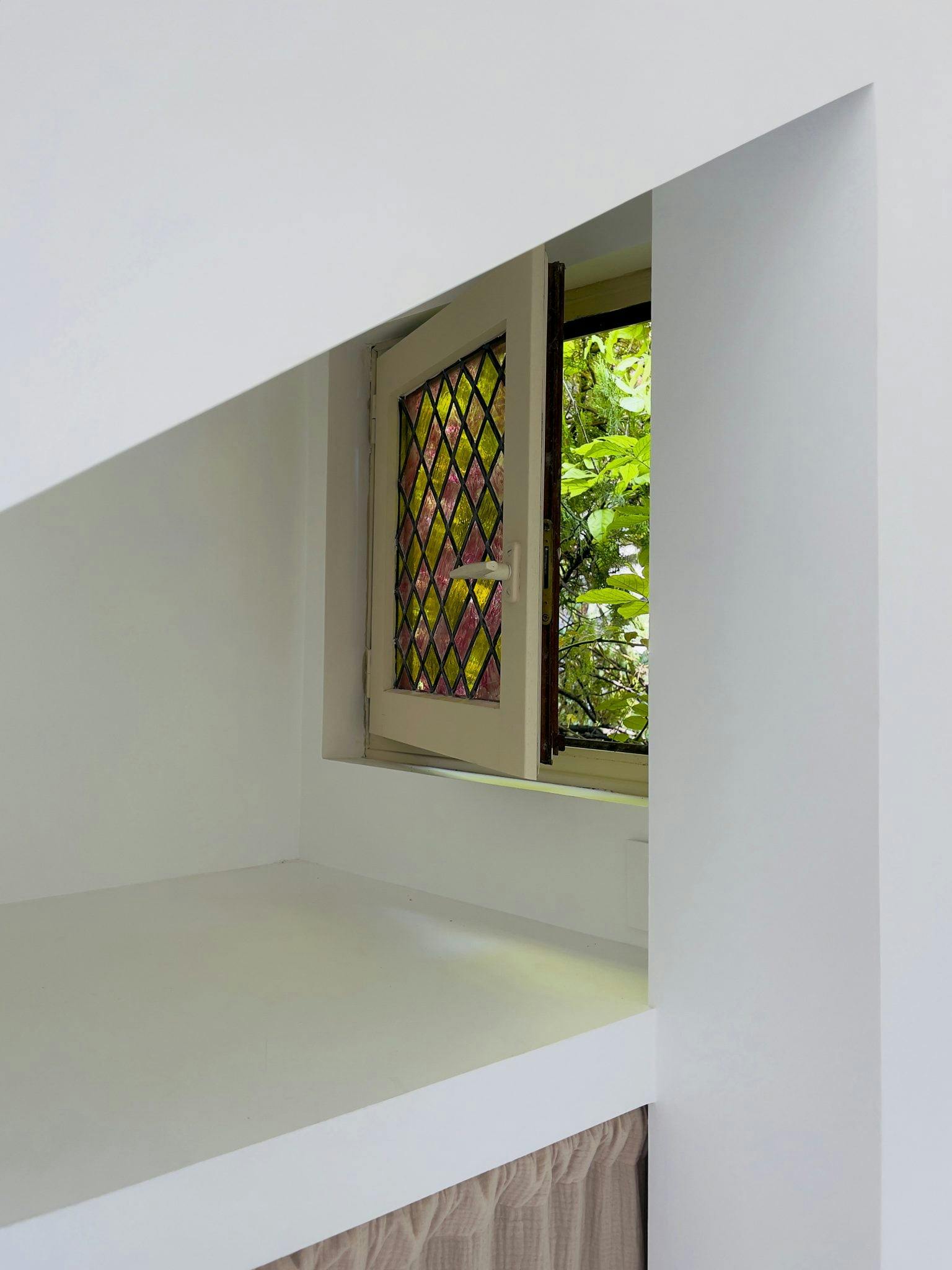 A small window with a diamond-patterned stained glass pane is set into a white wall. Outside, green foliage is visible through the window. The interior space features minimalist white surfaces with a beige curtain below.