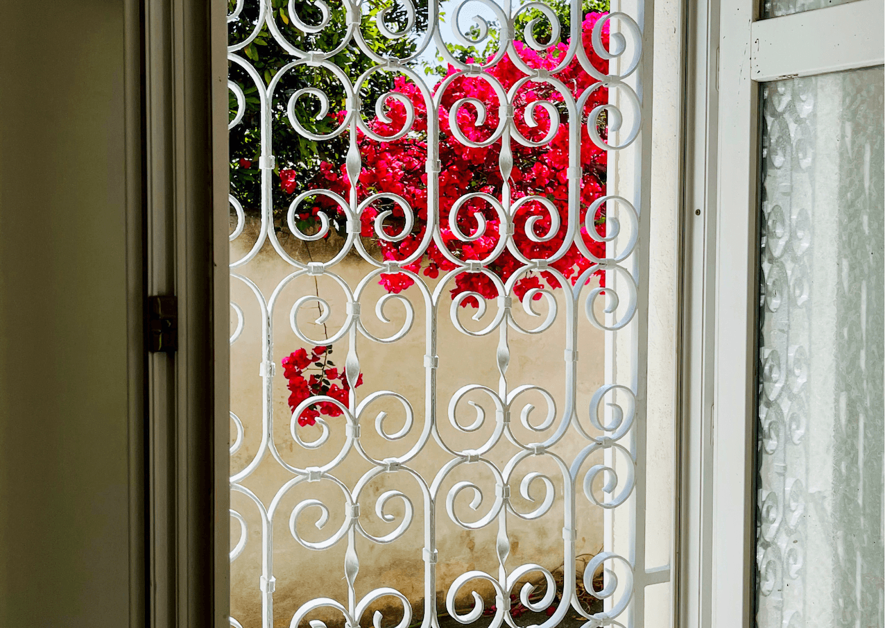 Une fenêtre ouverte avec des barreaux en fer forgé blancs et ornés offre une vue sur un mur de pierre orné de fleurs de bougainvilliers rouges vifs. Le cadre de la fenêtre et la ferronnerie complexe offrent un premier plan décoratif à l'arrière-plan luxuriant.