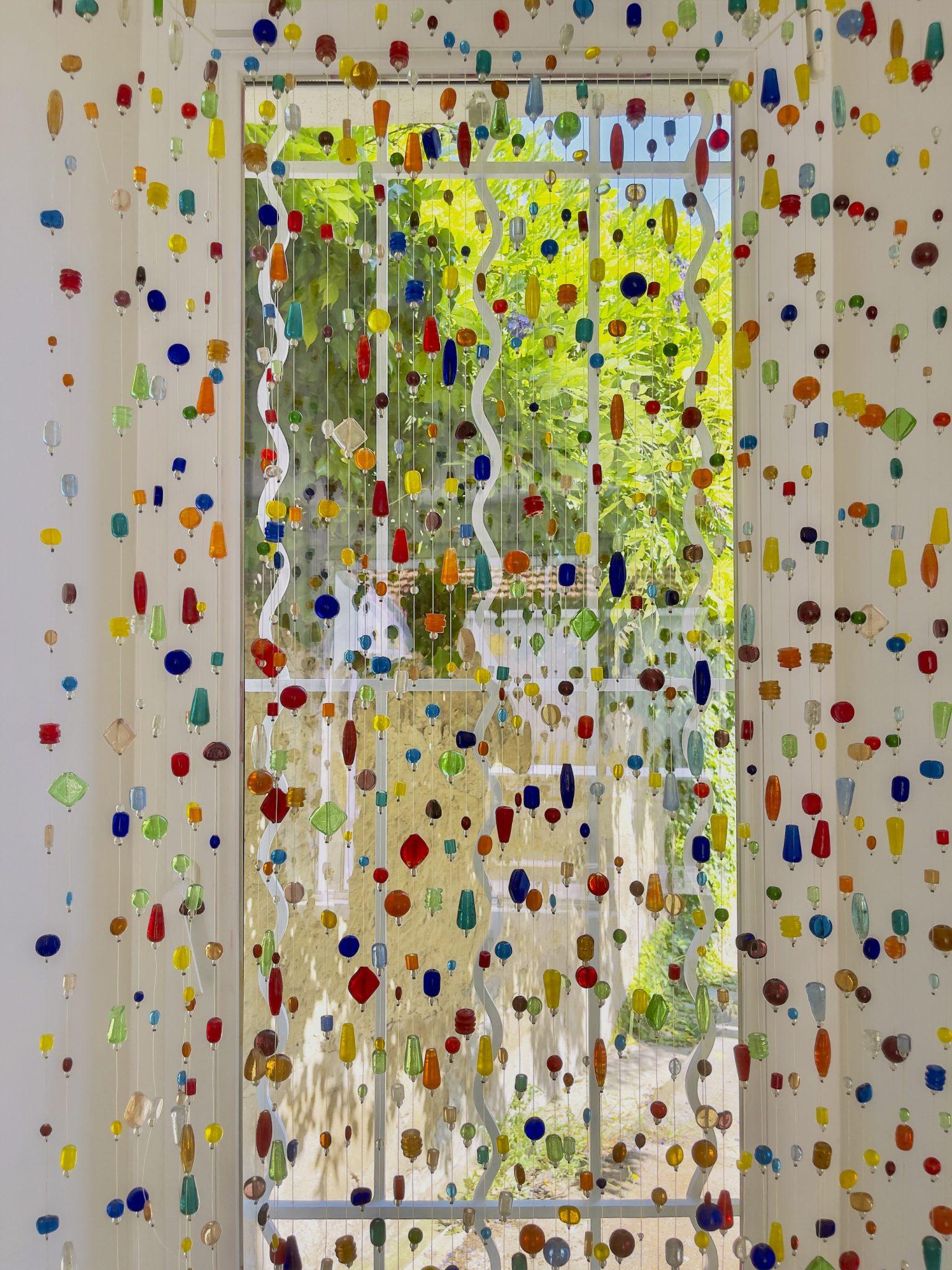 A window adorned with an array of hanging colorful beads and glass ornaments. The decorative pieces are suspended from multiple strings, allowing light to pass through and creating a vibrant and playful atmosphere. Greenery is visible through the window.