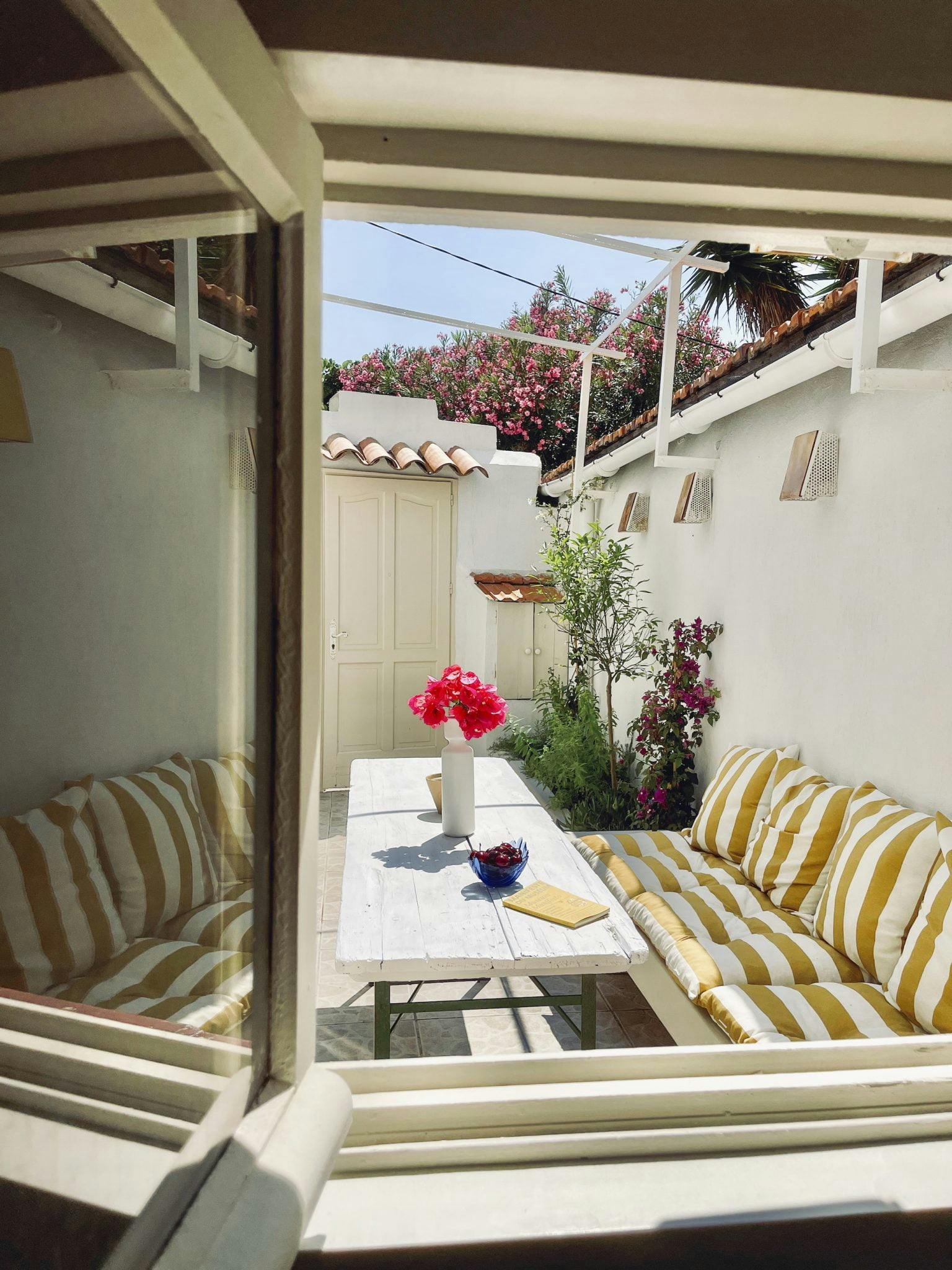 A cozy outdoor seating area viewed through a window, with yellow and white striped cushions on benches along a long wooden table. The table holds a vase of red flowers and a fruit bowl. The space is surrounded by white walls and adorned with flowering plants.