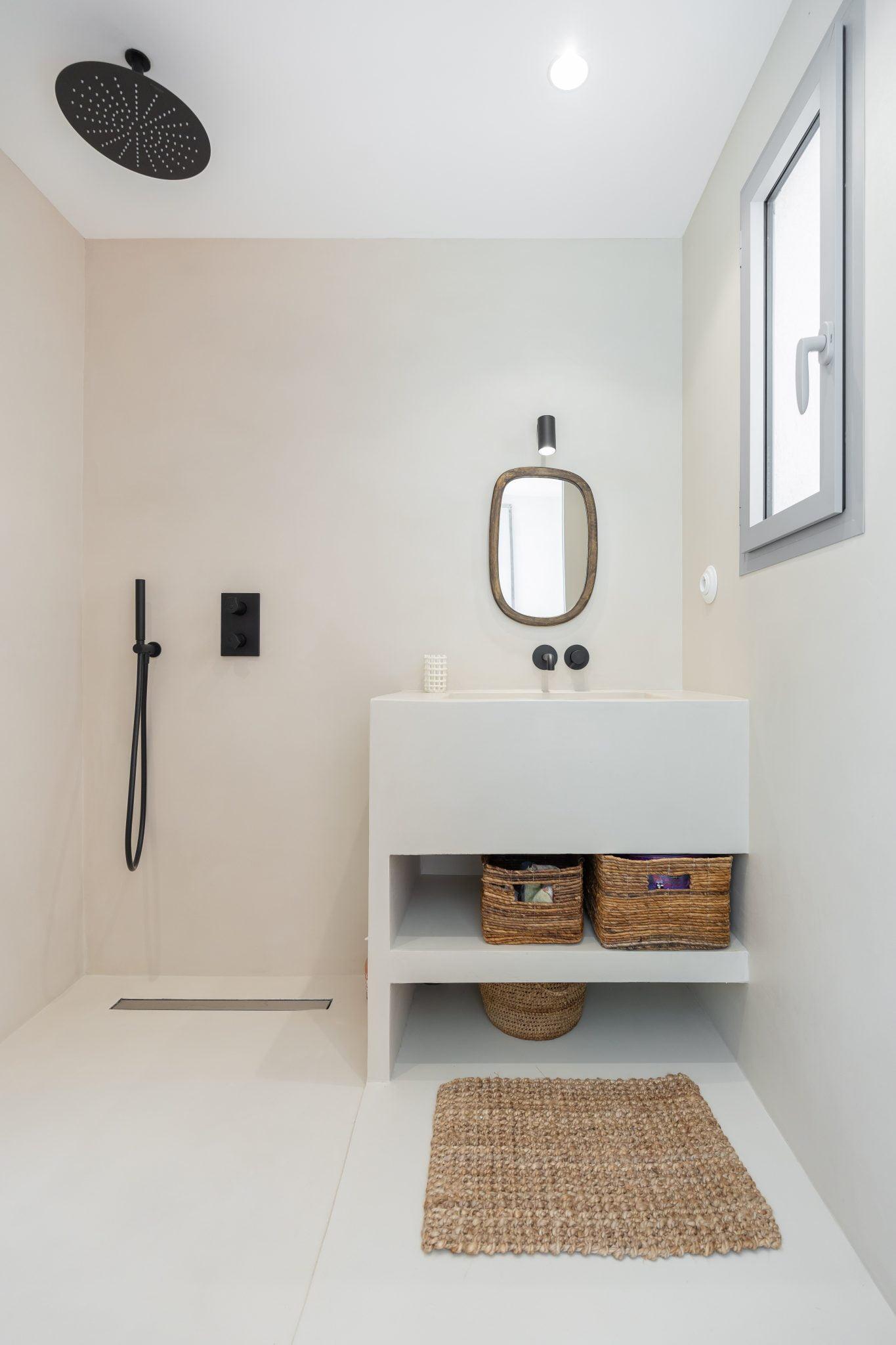 A modern and minimalist bathroom featuring a walk-in shower with a black rain showerhead and faucet. It includes a rectangular white sink topped with a circular mirror, two woven baskets on a shelf under the sink, a small wicker basket on the floor, and a window.