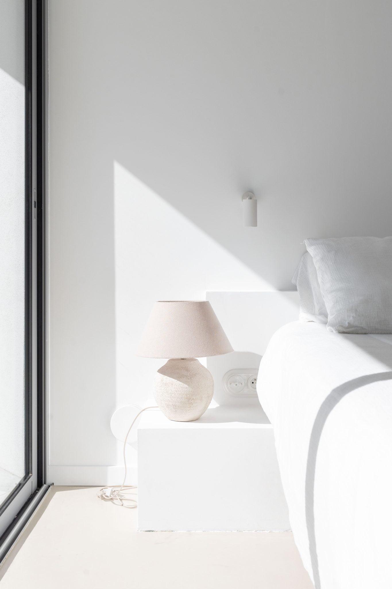 A minimalist bedroom with white walls and bright natural light streaming through a large window. The corner of a white bed and a bedside table with a beige lamp are visible. The design is simple and modern, with clean lines and light colors dominating the space.