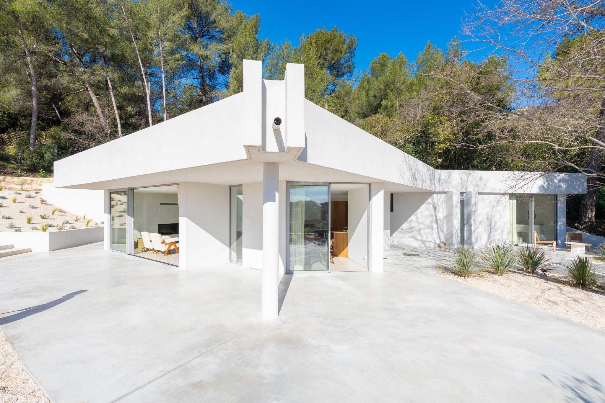 A modern and minimalist house with white walls and large glass windows is located in a sunny, wooded neighborhood. The house features angular architecture with a flat roof and a spacious outdoor patio surrounded by tall trees and a clear blue sky.