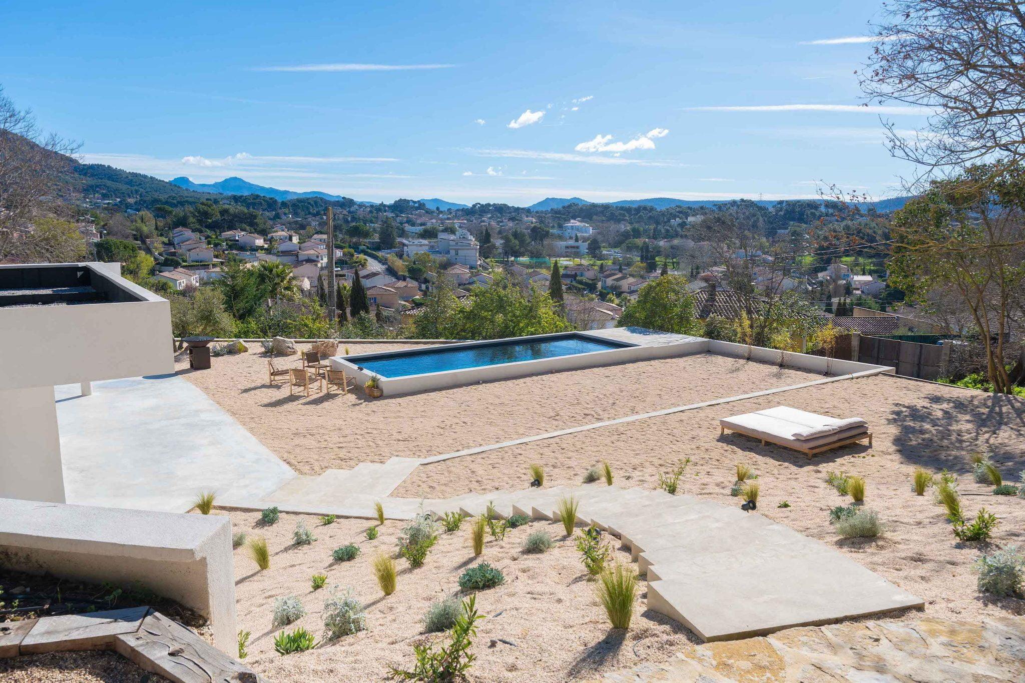 Une cour paysagée comprend une piscine rectangulaire avec une vue lointaine sur les collines et un ciel dégagé. Il y a des chaises longues et un petit coin salon autour de la piscine. Les sentiers et la verdure sont intégrés au terrain sablonneux pour une ambiance sereine et pittoresque.