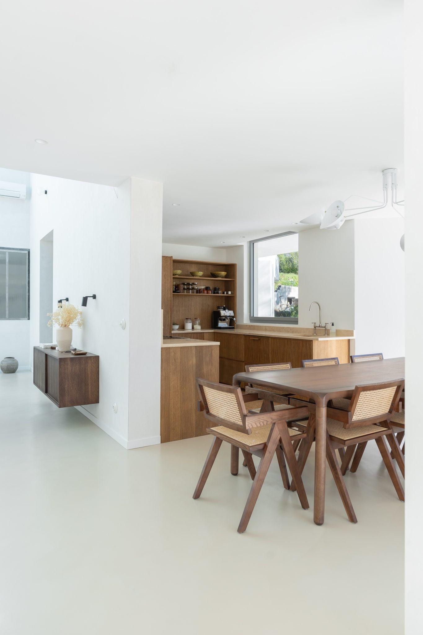 A minimalist and modern kitchen and dining area with clean white walls. The kitchen features wooden cabinets and open shelves, while the dining area includes a wooden table and four woven-back chairs. The space is illuminated by natural light from large windows.