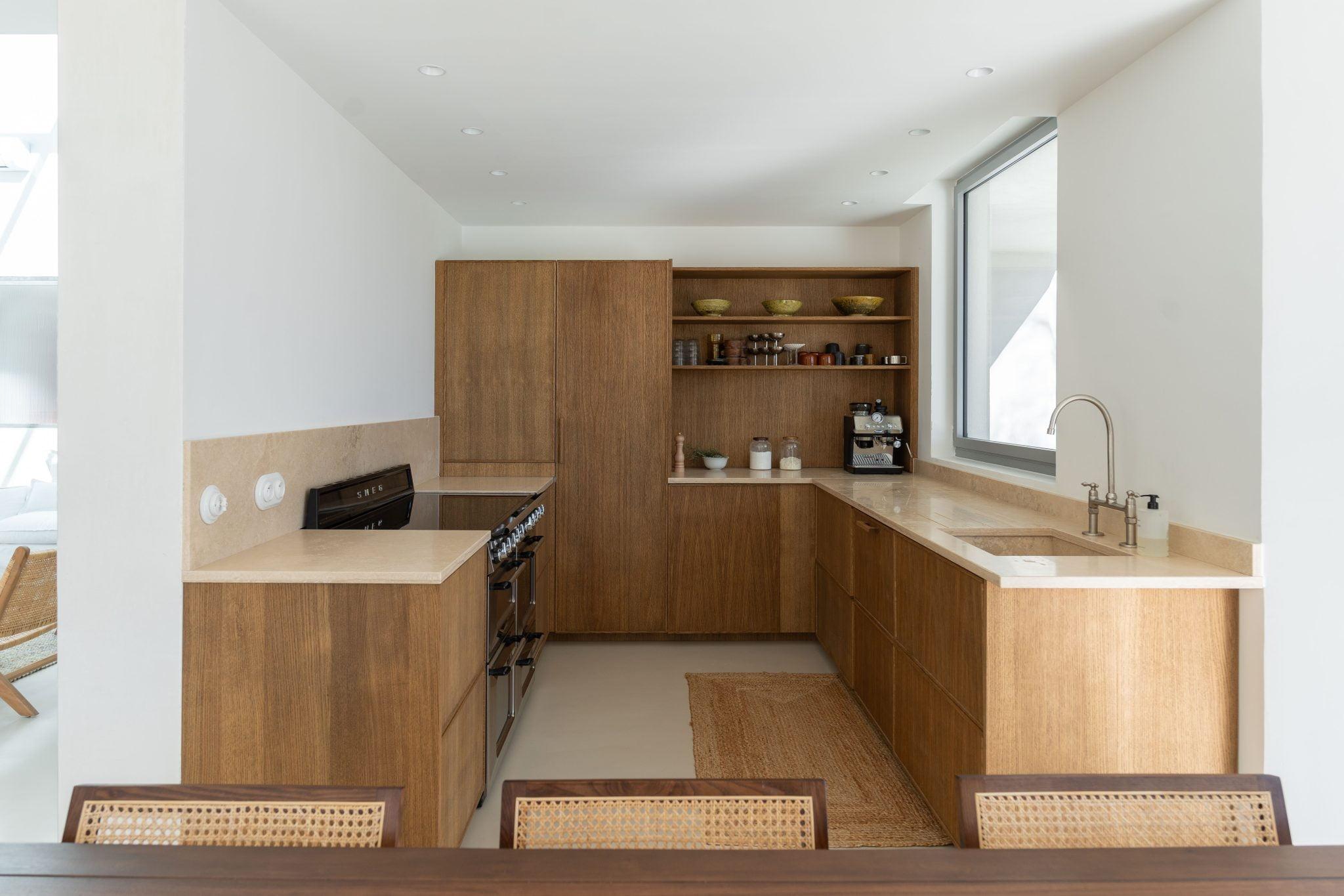 A modern kitchen featuring wooden cabinets and beige countertops. The kitchen includes a gas stove, an oven, open shelves with bowls and jars, a coffee maker on the counter, a sink with a minimalist faucet, and a large window that lets in natural light.