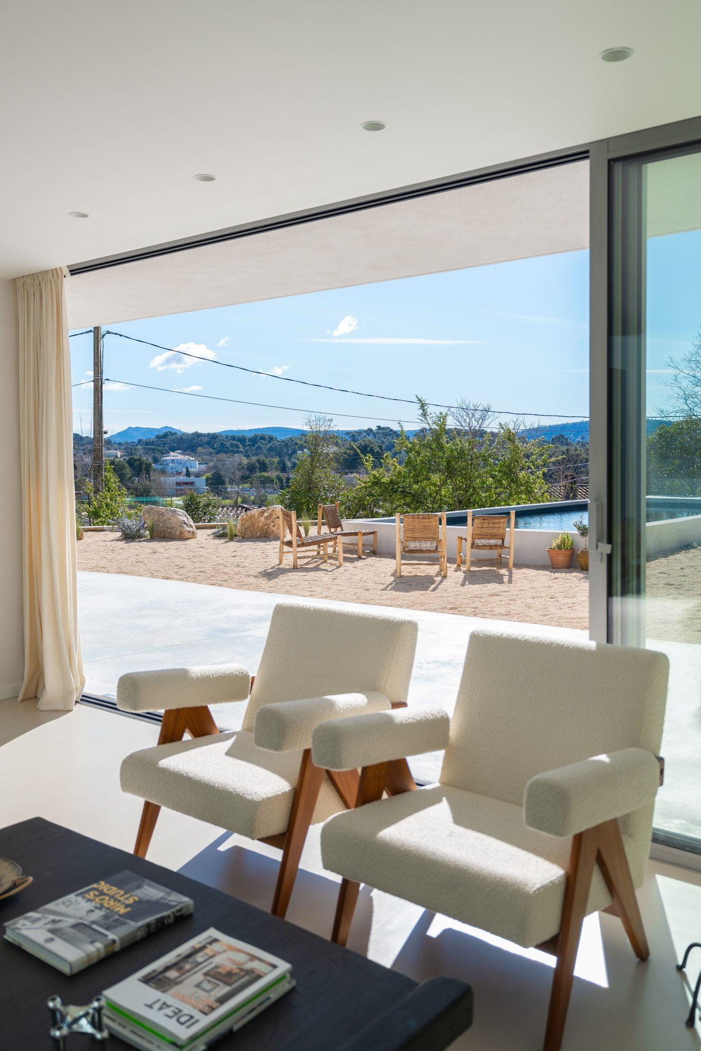 Un salon moderne avec deux fauteuils blancs et une table avec des magazines. Les grandes portes coulissantes en verre offrent une vue imprenable sur un patio extérieur ensoleillé, des chaises en bois et un paysage pittoresque avec des collines, des arbres et un ciel bleu clair.