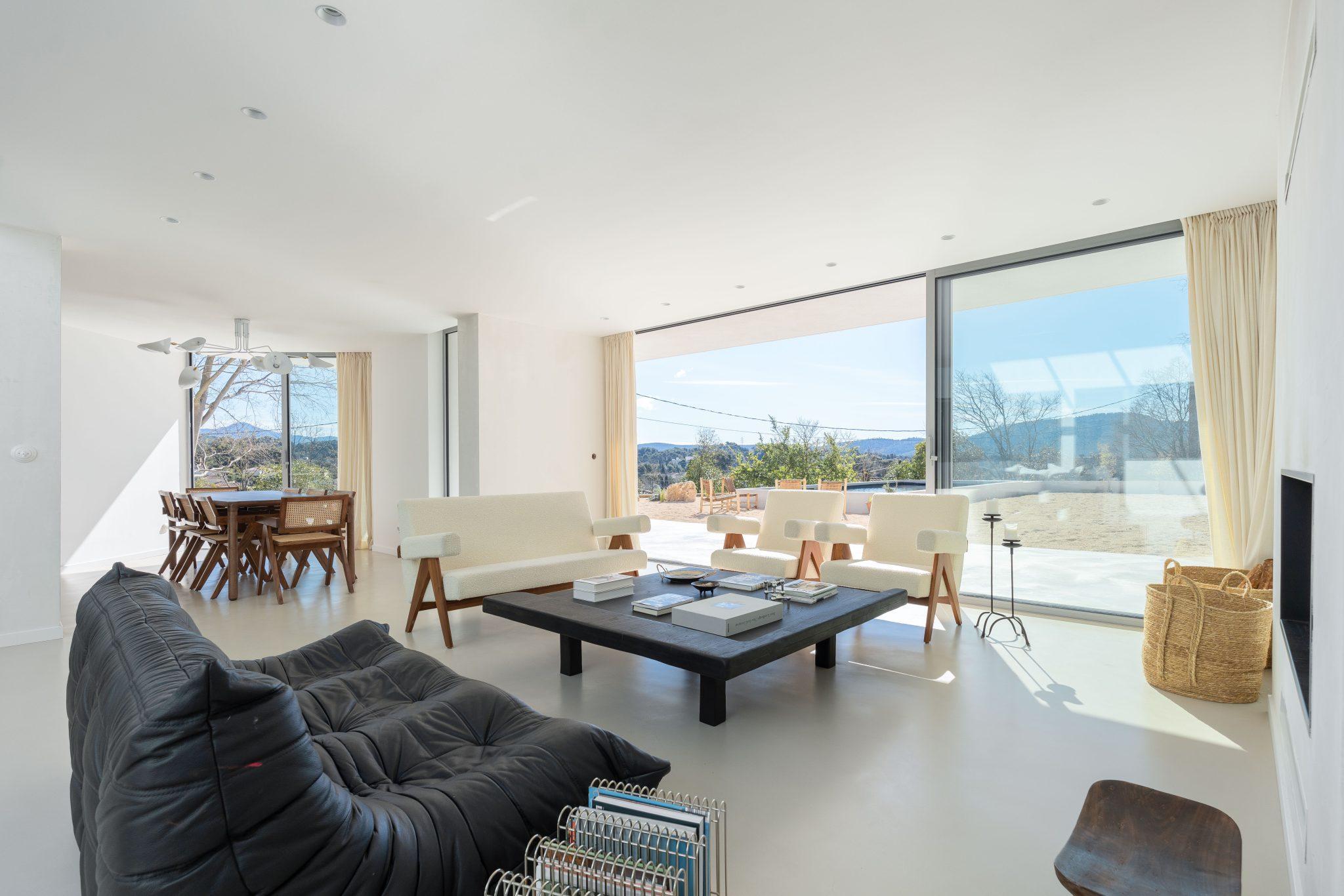 A modern and spacious living room featuring a black sofa, wooden chairs with white cushions, and a large coffee table with books and magazines. Large bay windows provide a panoramic view of the outdoors, and a dining area is visible in the background.