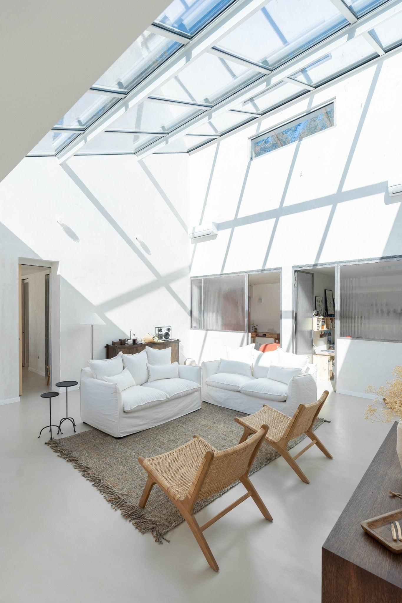 A spacious and sunny living room with large skylights. The room features white walls, a plush white sofa, a matching loveseat, and two woven wood chairs. A textured rug covers the floor, and a sideboard with decorative items is on the right. Doors are visible in the background.