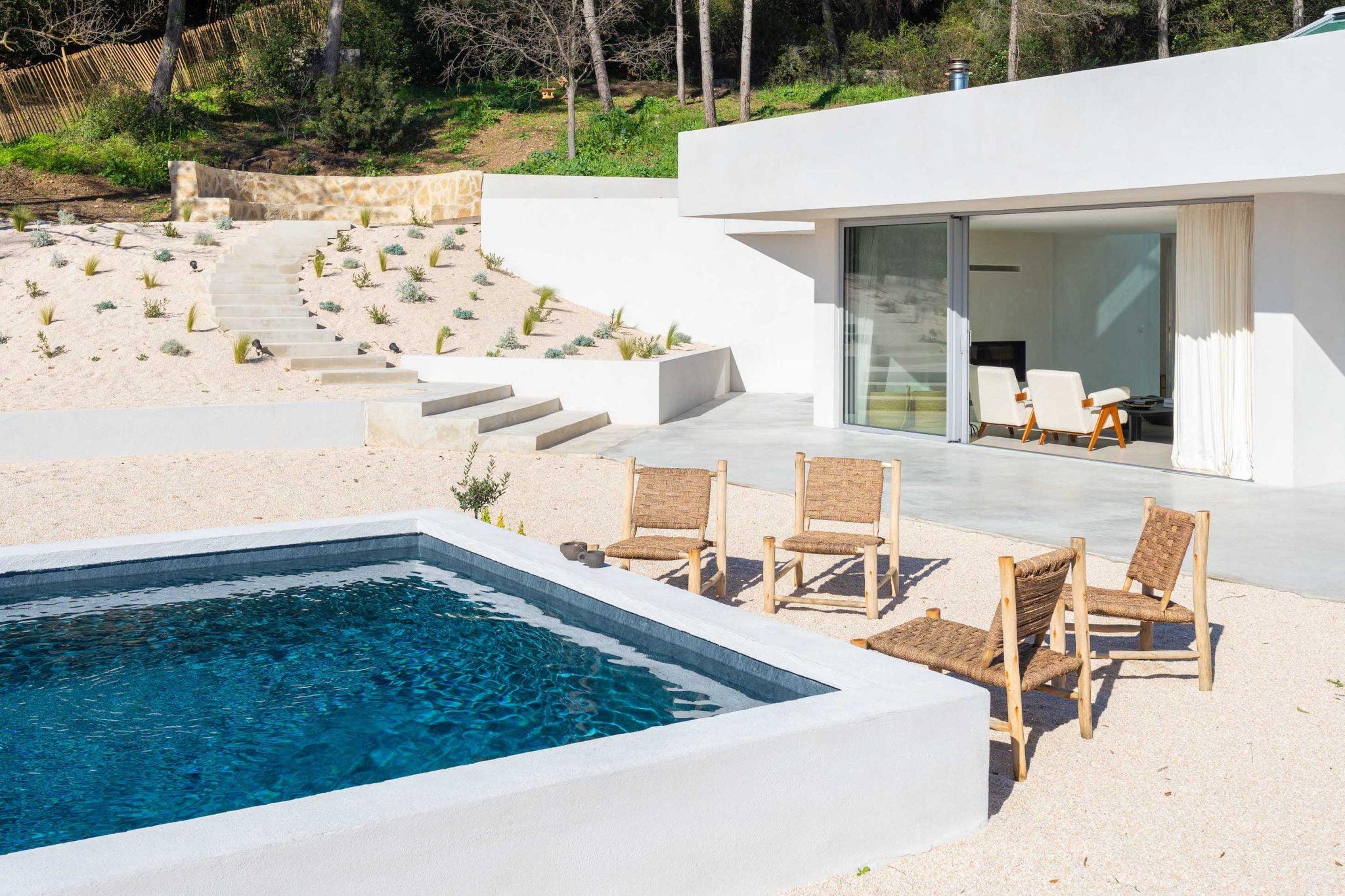 Une maison blanche moderne dispose d'un patio extérieur avec quatre chaises longues en bois et une piscine rectangulaire. Le patio est entouré d'un terrain sablonneux avec des marches menant à un espace paysager avec de petits arbustes et des murs de soutènement en pierre. Des arbres sont visibles en arrière-plan.