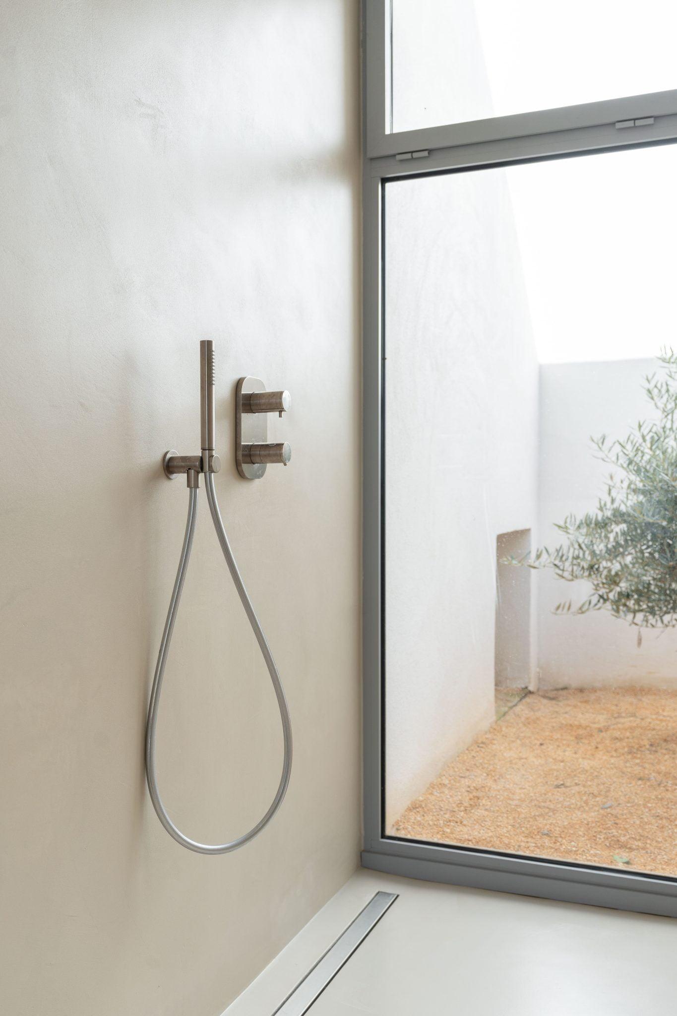 Salle de bains minimaliste avec commandes de douche murales et pomme de douche amovible à côté d'une grande fenêtre en verre transparent. La fenêtre révèle un petit espace extérieur avec des murs blancs et un olivier. Le design général est moderne et épuré.