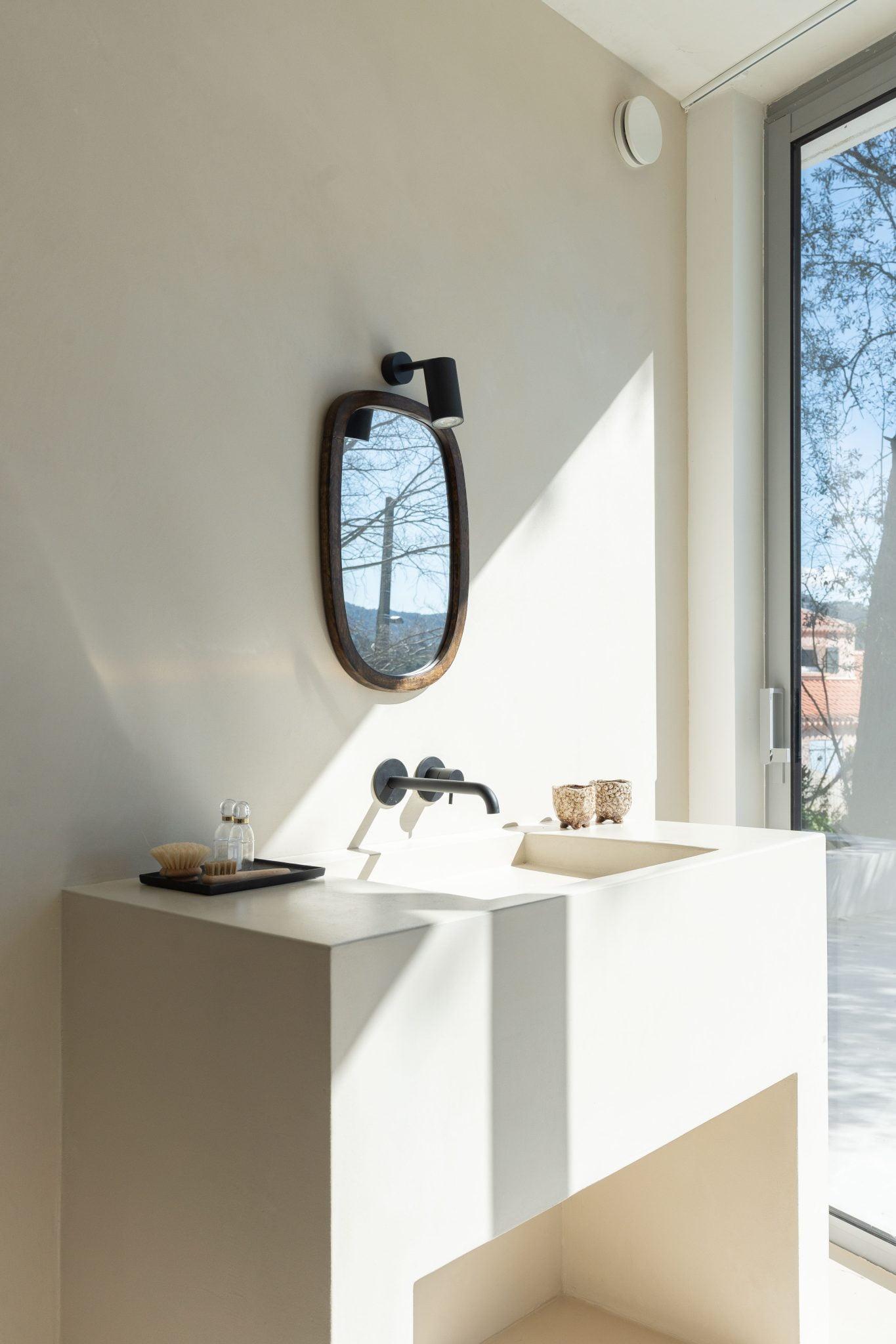 A minimalist bathroom featuring a rectangular sink with a wall-mounted faucet. Above the sink is an oval mirror with a black frame and a light fixture. The sink countertop has a black tray with soap and a small bottle. A large window allows natural light to fill the space.