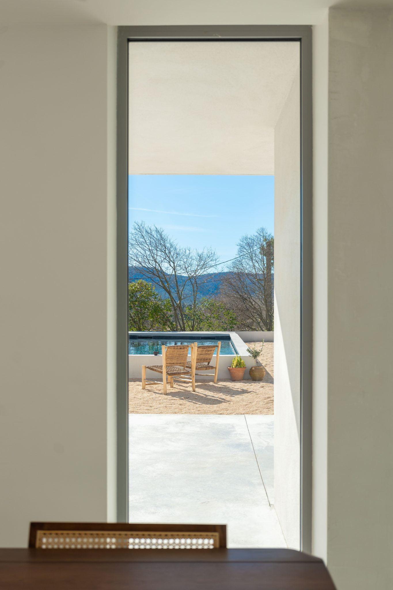 A view through a tall, narrow window reveals an outdoor patio with two wooden chairs facing a rectangular pool. Beyond the pool, trees and a clear blue sky with distant mountains are visible on a sunny day. In the foreground, part of a wooden table can be seen inside.