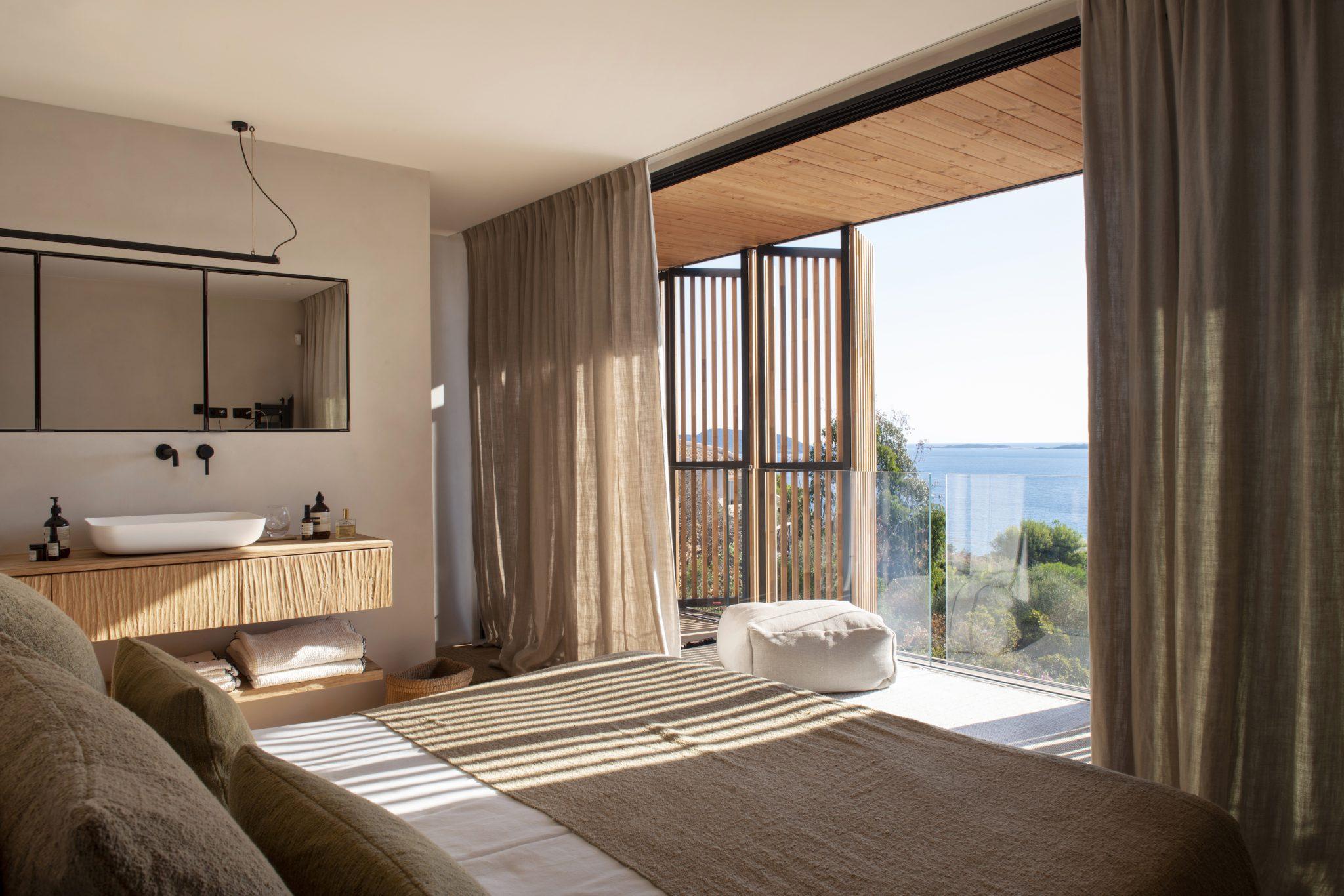 A modern bedroom with large glass doors opening onto a balcony overlooking a body of water. The room features neutral tones, with a bed dressed in green and beige linens, a wooden headboard, and a floating sink with a mirror. Soft light filters through sheer curtains, creating a tranquil atmosphere.