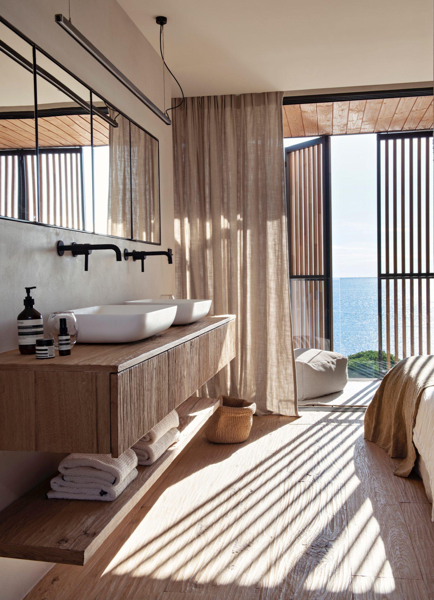 A modern bathroom with large windows offering an ocean view. It features a wooden countertop with two white sinks, a large rectangular mirror, and neatly folded towels on an open shelf below. Light filters through beige curtains.