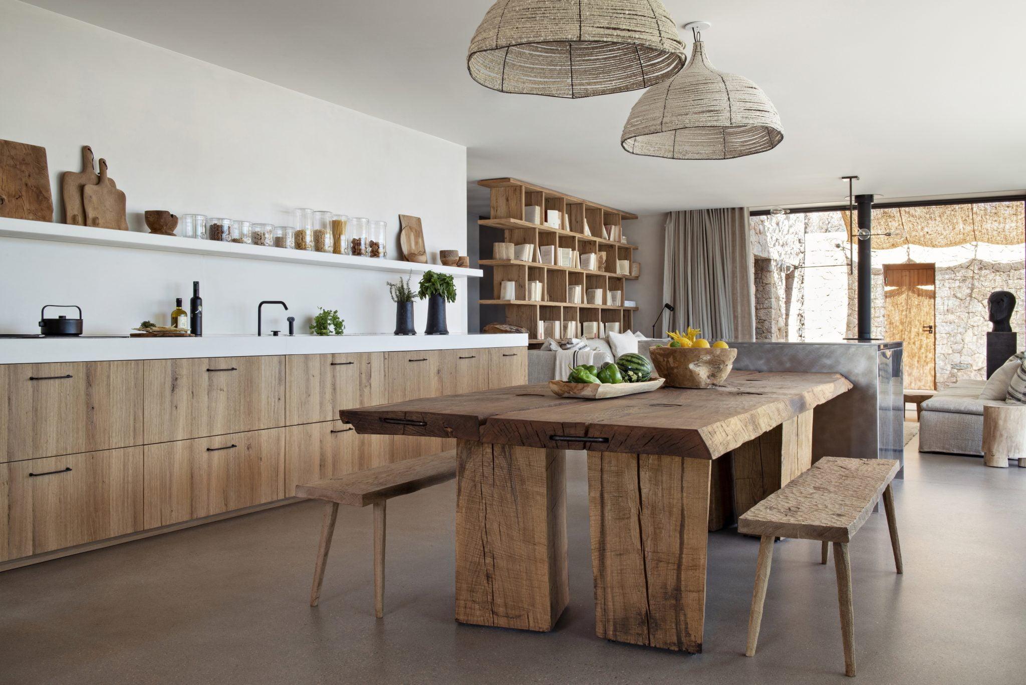 A spacious and modern kitchen with a rustic touch, featuring wooden cabinets, a large wooden dining table, and benches. Two woven pendant lights hang above. In the background, shelves filled with various items are visible, and large windows let in natural light.