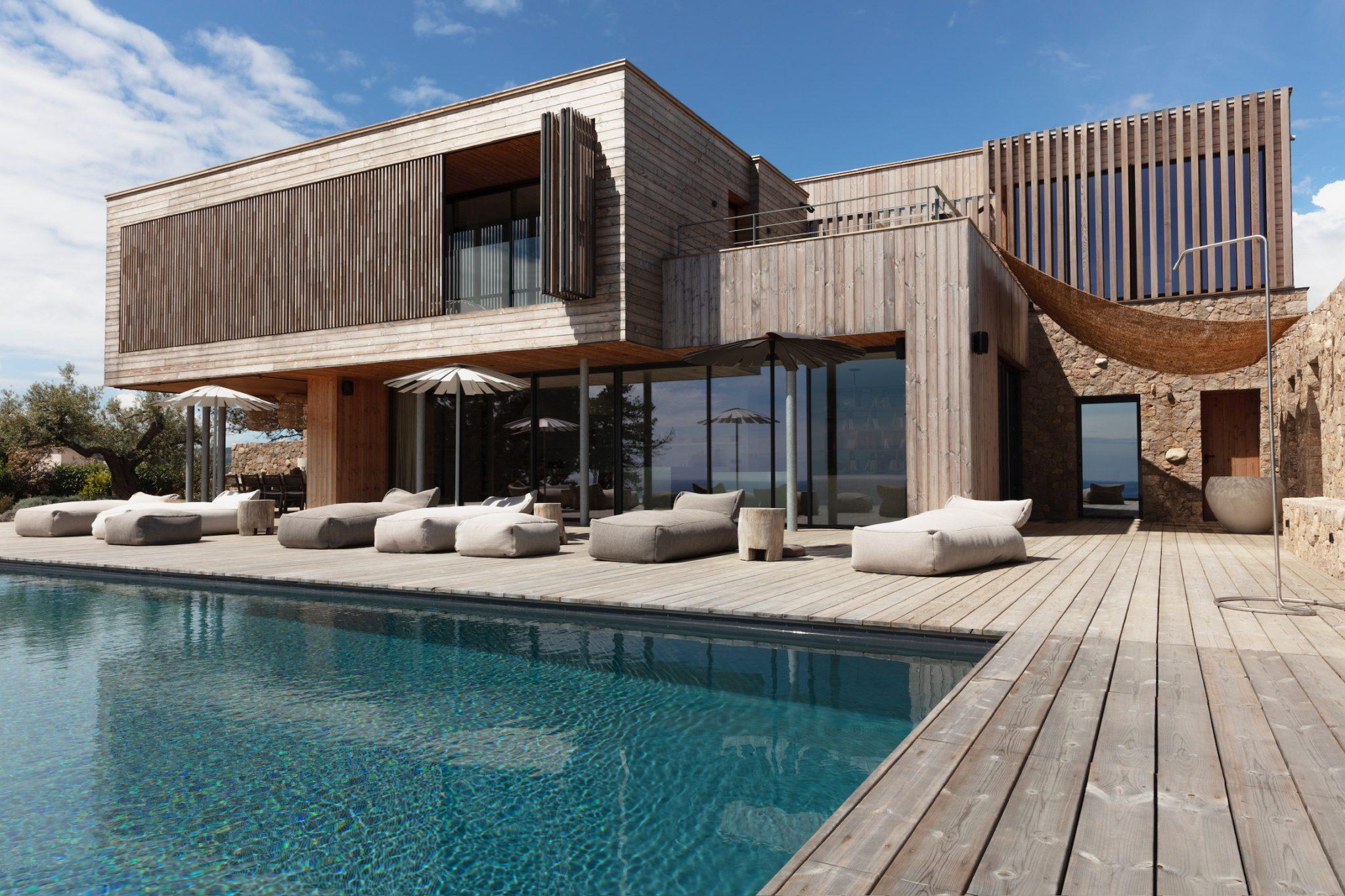 A modern two-story wooden house with large glass windows, featuring a spacious wooden terrace and an infinity pool. The terrace is furnished with several cushioned lounge chairs and umbrellas, with an ocean view in the background under a partly cloudy sky.