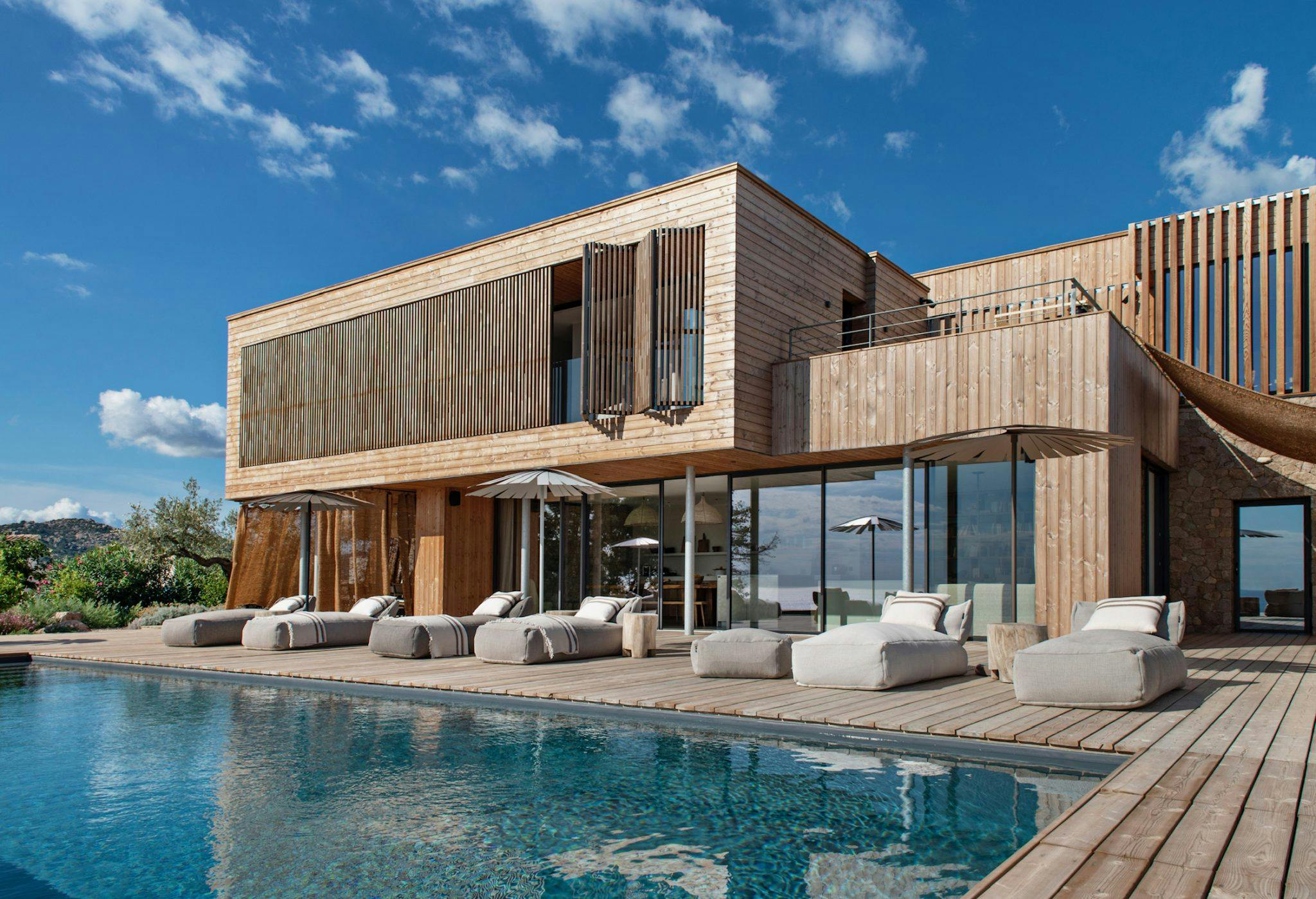 Une maison en bois moderne à deux étages avec de grandes baies vitrées et des zones ombragées donne sur une piscine avec des chaises longues rembourrées alignées sur la terrasse en bois. Le ciel est clair avec quelques nuages, et la verdure entoure la zone.