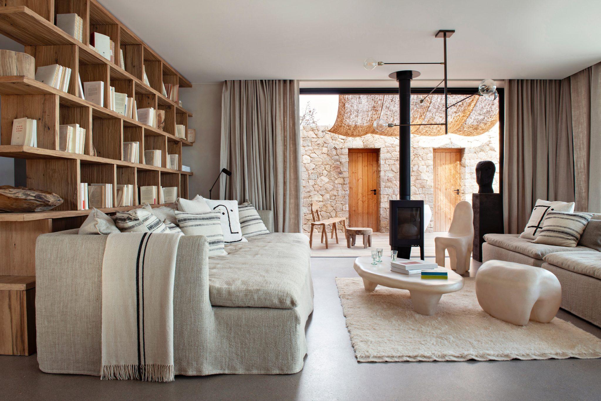 A cozy living room features a beige corner sofa, wooden shelves filled with books, an eclectic white coffee table, and a modern wood stove. Natural light filters through large windows dressed in bright curtains, leading out to a stone outdoor patio.