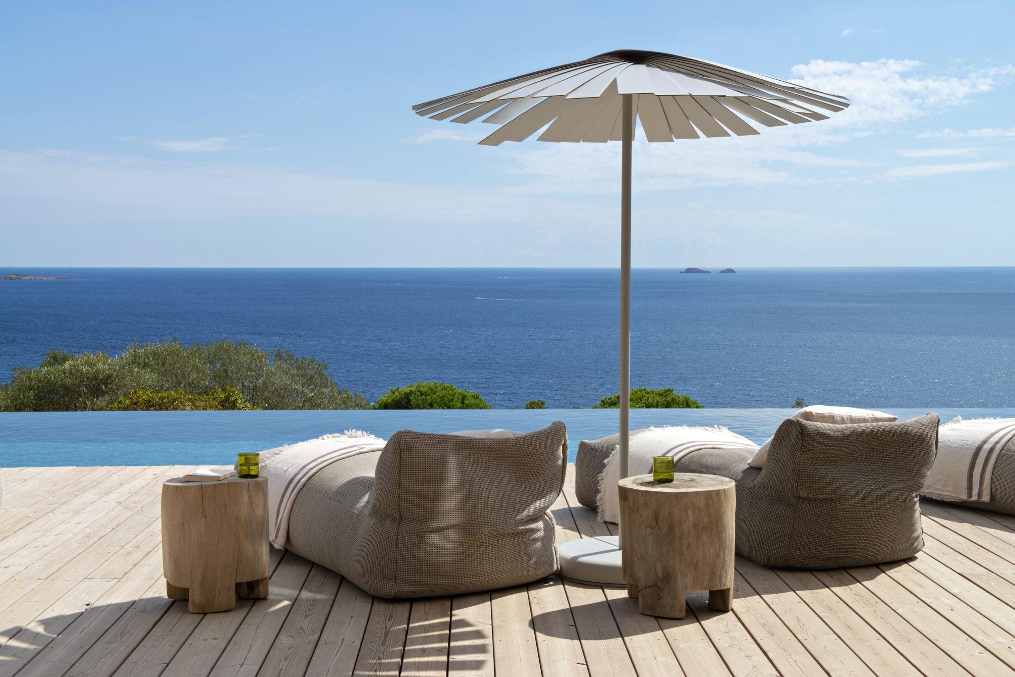 Un aménagement extérieur serein avec des chaises longues moelleuses et des tables d'appoint en bois sur une terrasse en bois surplombant un océan calme et vaste. Un parasol au design moderne fournit de l'ombre. Le ciel est clair avec un soupçon de nuages, et de petites îles sont visibles au loin.