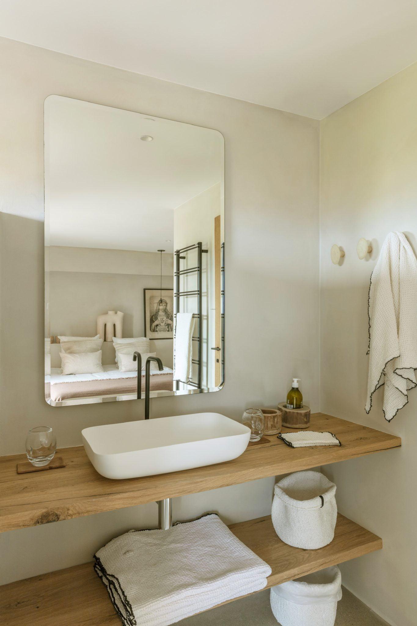 A minimalist bathroom featuring a large mirror, a modern white sink, and a sleek faucet on a wooden countertop. Below the countertop, towels and neatly organized baskets are arranged. In the background, a serene bedroom with a neatly made bed is visible through an open door.