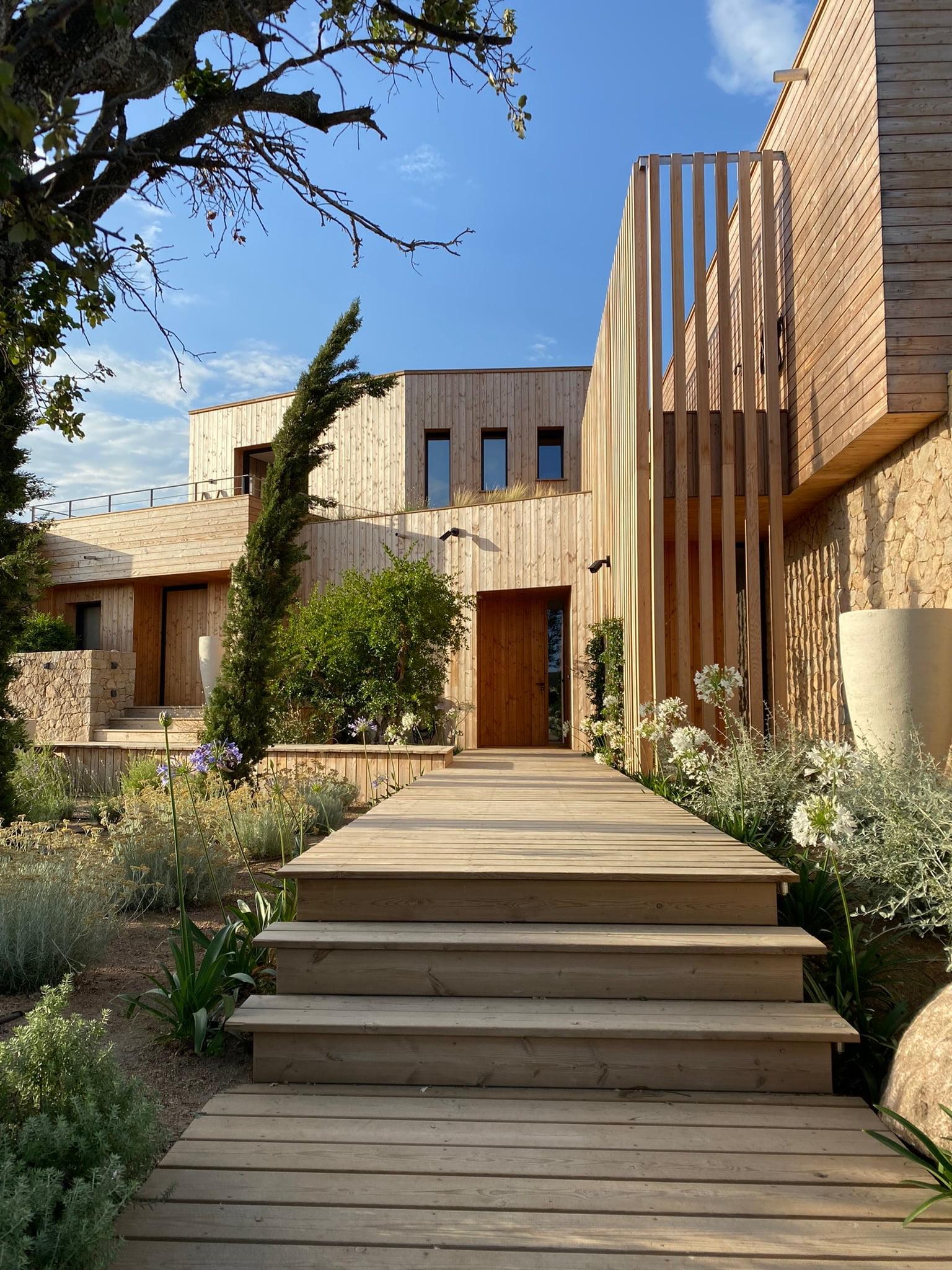 Une maison en bois moderne au design minimaliste entourée de verdure. L'entrée principale comporte un escalier menant à un chemin en bois flanqué de plantes. La façade combine des planches de bois horizontales et des accents de pierre, avec de grandes lattes verticales sur le côté droit.