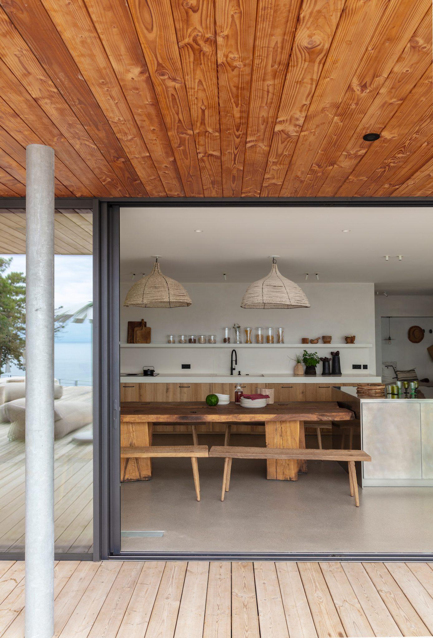 View of a modern kitchen and dining area through a sliding glass door. The space features wooden ceilings, a long wooden dining table with benches, wicker light fixtures, and white countertops. Outside, a wooden deck and a glimpse of the ocean are visible.