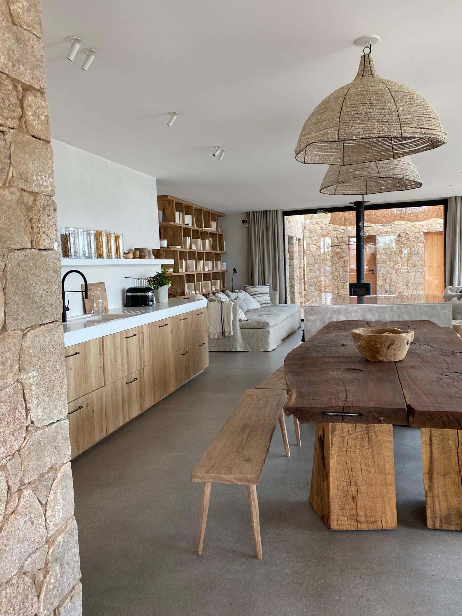 A modern and minimalist kitchen and dining area with wooden furniture. The kitchen features light wood cabinets and open shelving, while the dining table is paired with benches. Large wicker pendant lights hang from the ceiling, and a cozy living room is visible in the background.