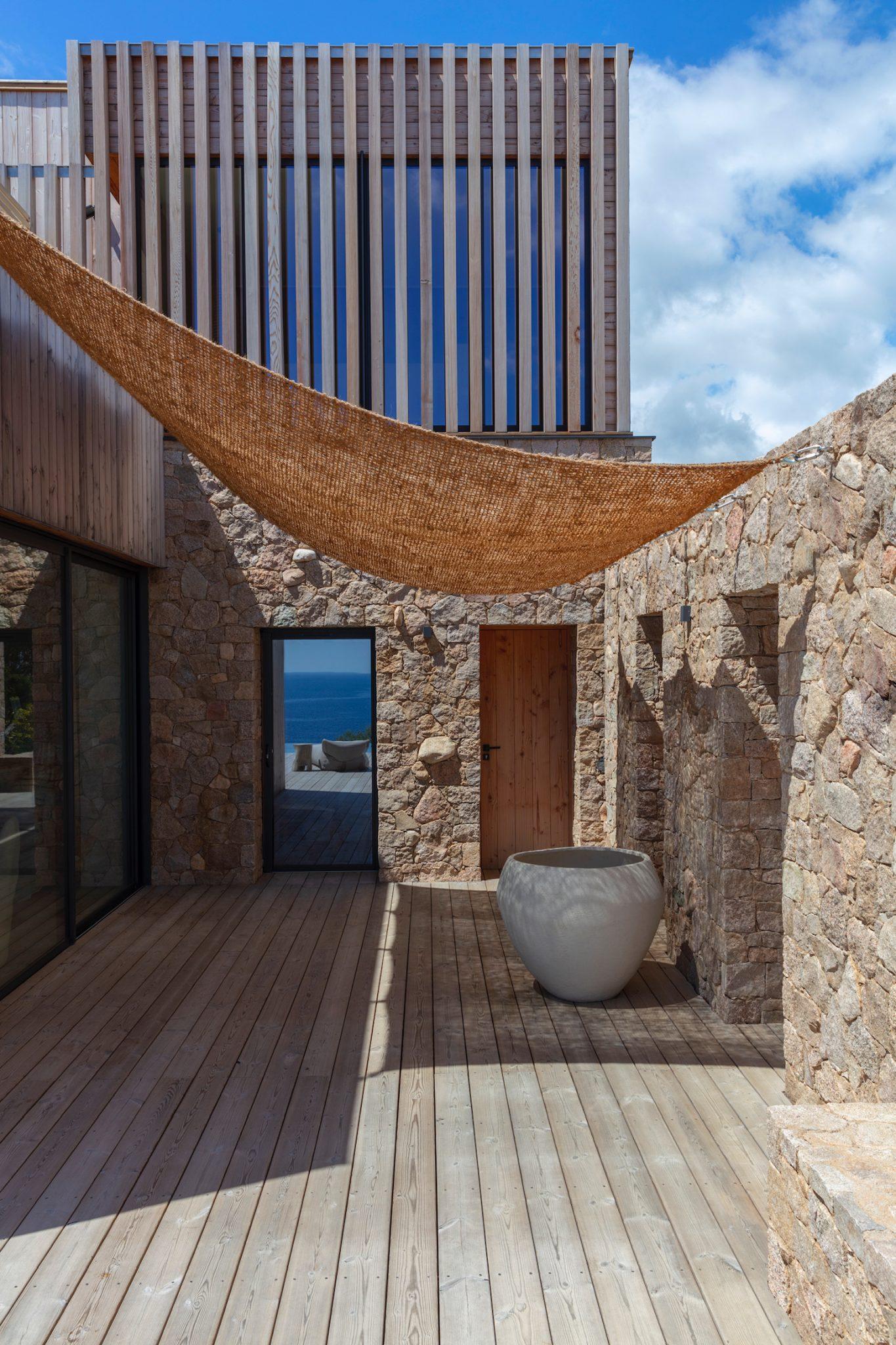 A modern terrace with stone walls and light wood decking, featuring a large round planter and a wooden door. A brown shade sail is stretched overhead. In the background, an opening reveals a partial view of the ocean and the blue sky.