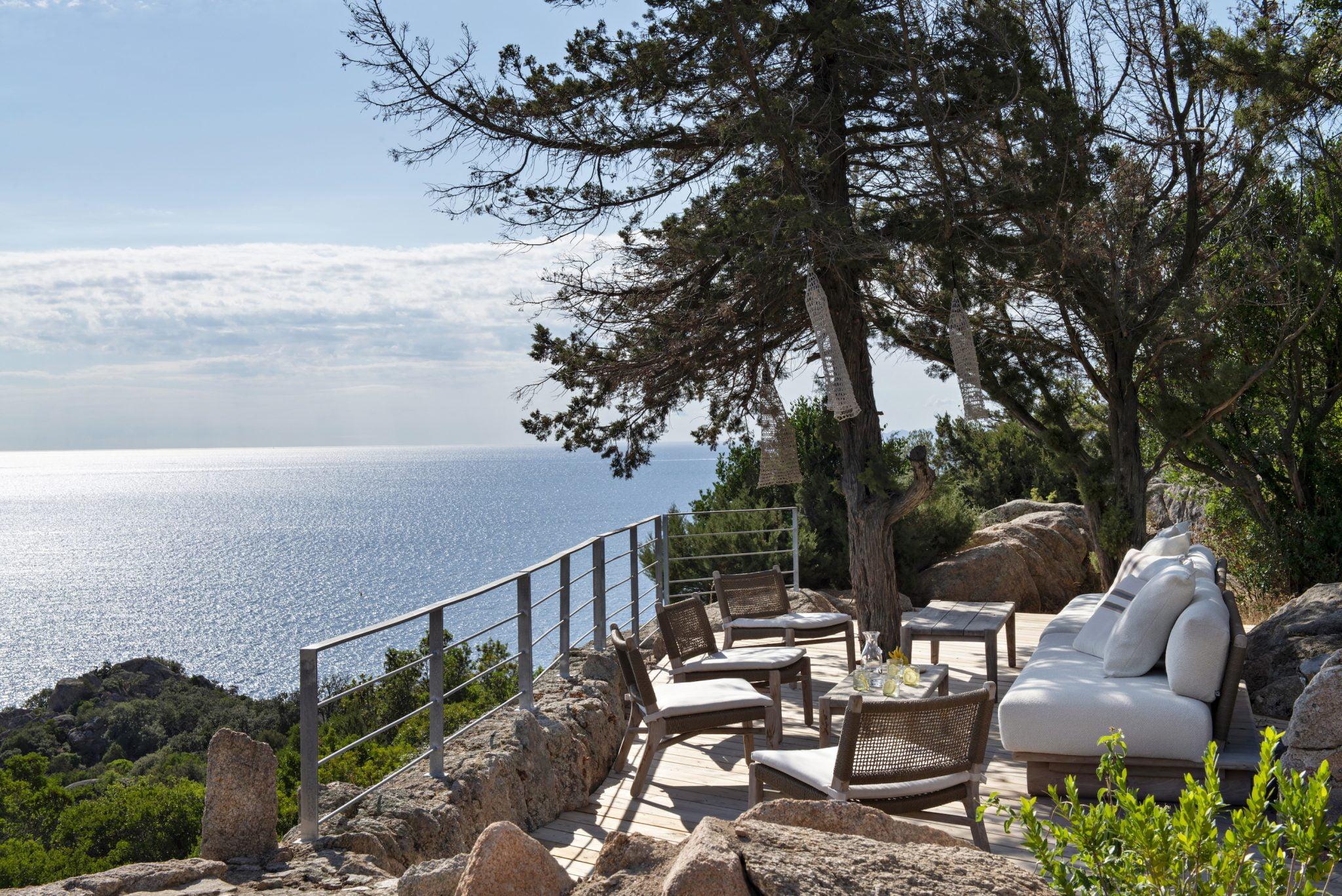 Une terrasse extérieure pittoresque avec des meubles en bois, notamment des chaises rembourrées et une table avec des boissons, surplombe un océan serein. La zone est entourée d'arbres et de verdure, avec un ciel clair et ensoleillé en arrière-plan. Une balustrade en métal entoure la terrasse, améliorant la vue sur l'océan.