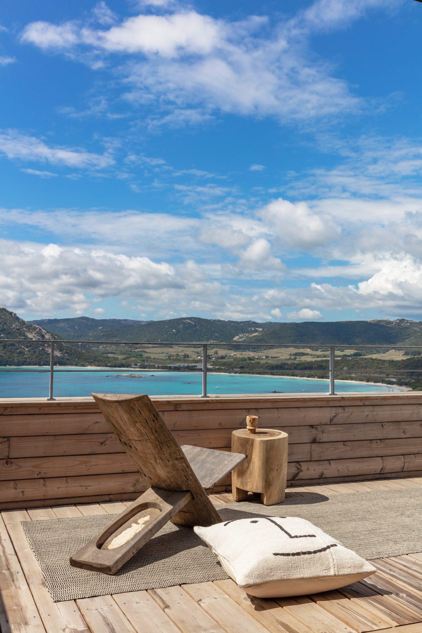 A rooftop wooden terrace featuring an elegant rustic wooden lounge chair and a cylindrical side table. A white cushion with a simple black design rests on a woven rug. The background showcases a breathtaking view of a blue ocean and lush green hills under a partly cloudy sky.