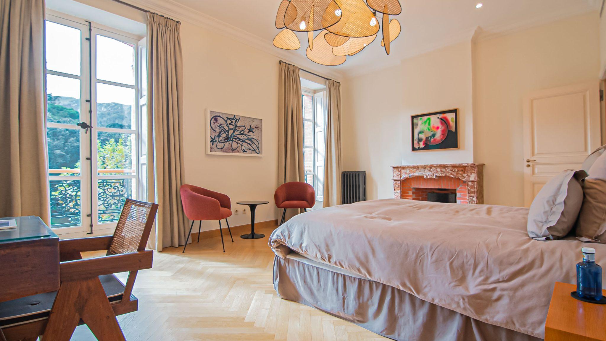 A spacious and bright bedroom with large windows and beige curtains. It includes a double bed, a wooden desk with a chair, two red armchairs with a small table, a brick fireplace adorned with artwork, and a unique pendant light. Natural light fills the room.