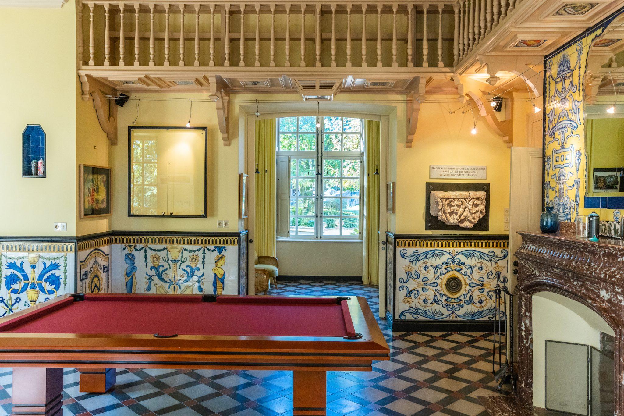 Une pièce avec une table de billard rouge au centre, décorée de carreaux bleus et jaunes complexes sur les murs. Il y a un coin salon confortable avec vue sur la fenêtre, une cheminée avec un manteau en marbre sur la droite et un loft en bois au-dessus. Diverses œuvres d'art ornent les murs.