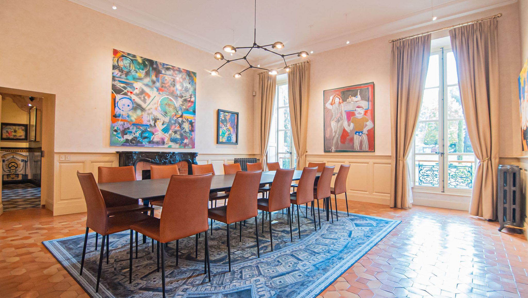 Une salle de conférence spacieuse et bien éclairée avec une longue table rectangulaire entourée de chaises orange. Les murs présentent des peintures abstraites et de grandes fenêtres avec des rideaux beiges permettent à la lumière naturelle de remplir la pièce. Un tapis à motifs complète le sol carrelé aux tons chauds.