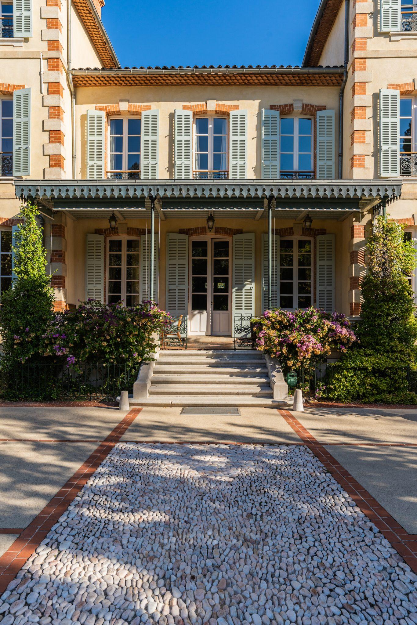 A large house with a beige exterior, green shutters, and a central porch with a black wrought-iron awning. It features a stone walkway flanked by lush greenery, flowering shrubs, and carefully trimmed hedges. The bright blue sky enhances the scene.