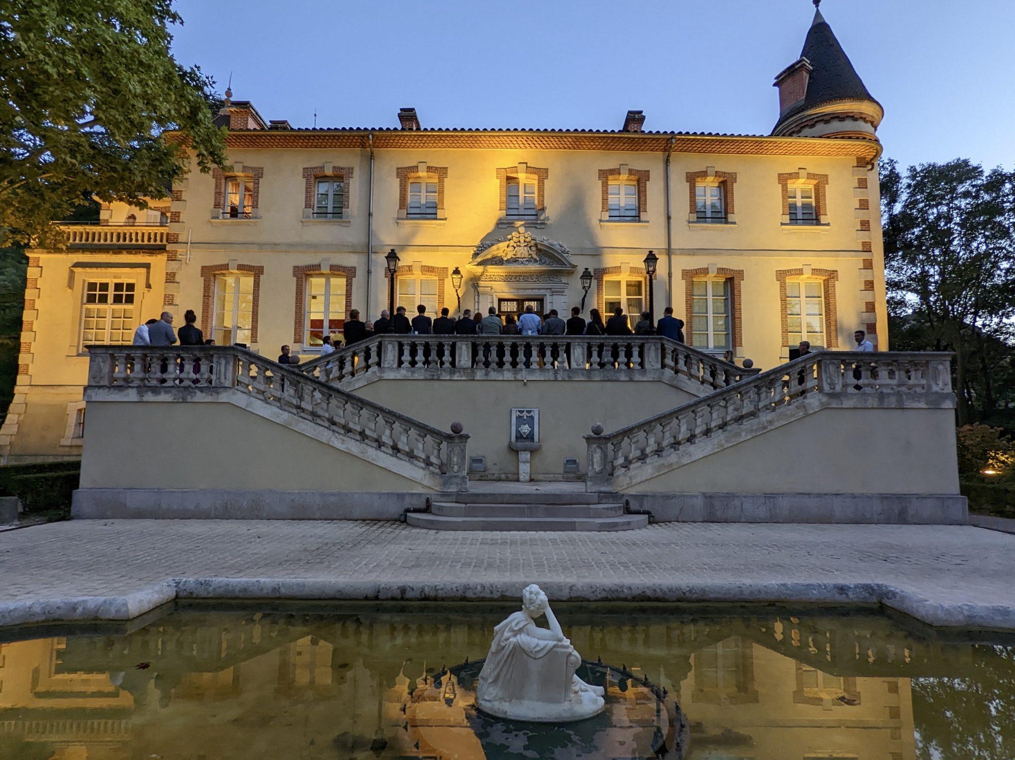 A large historic building with arched windows and a turret, illuminated in the evening. People are gathered on the balcony and stairs. A stone staircase leads to a rectangular reflecting pool with a white statue at its center. Trees frame both sides of the scene.