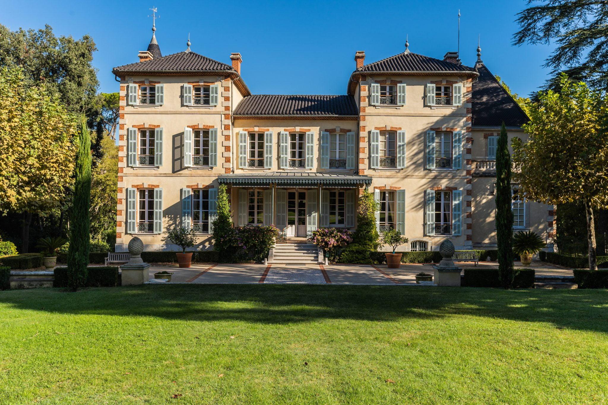 Un grand et élégant manoir avec des murs de couleur crème et des toits de tuiles sombres est entouré d'une pelouse verte luxuriante et de jardins bien entretenus. La maison dispose de nombreuses fenêtres avec volets et d'un porche central. Les arbres et les plantes en pot ajoutent à l'atmosphère sereine.