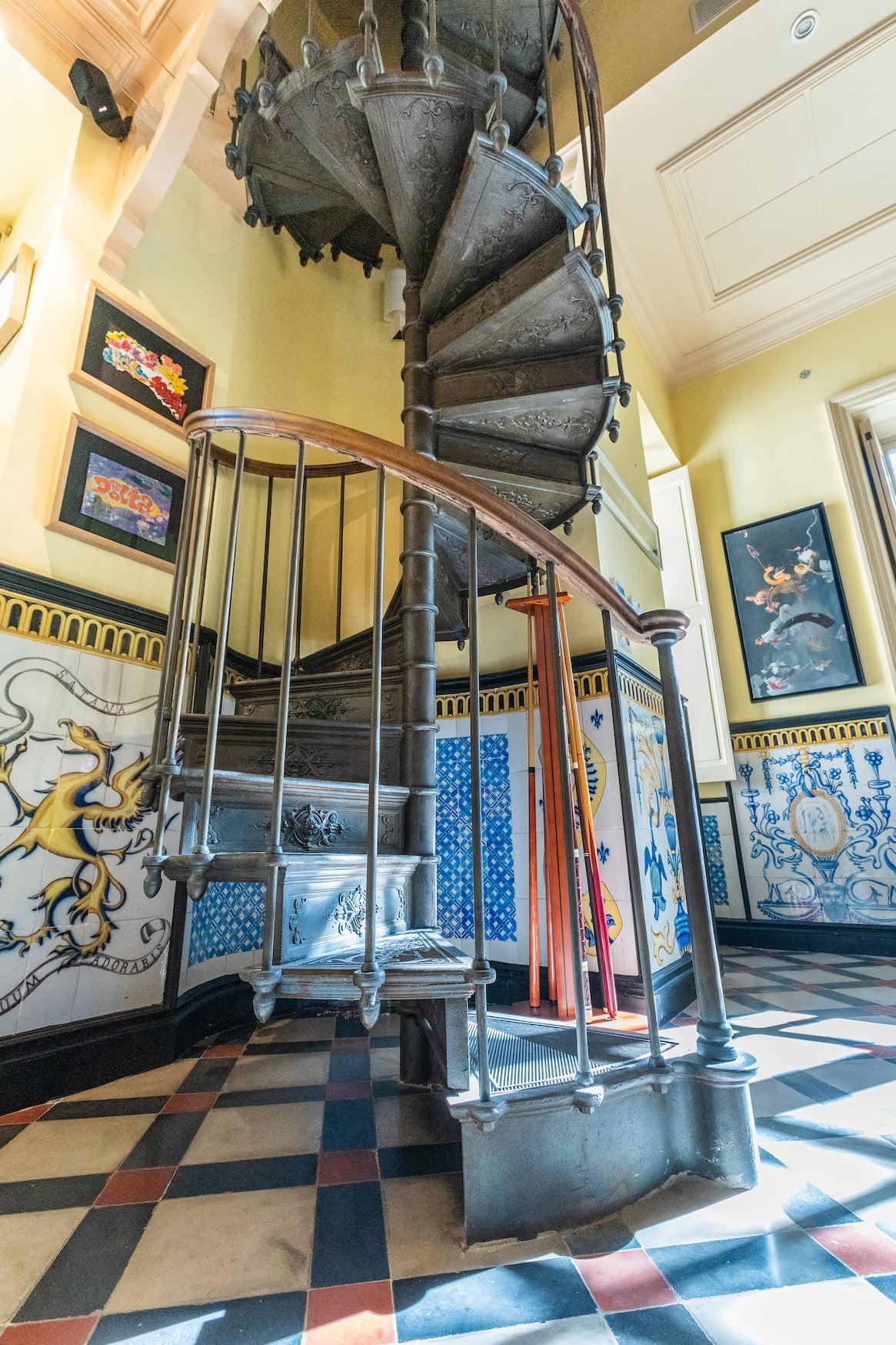 A complex spiral staircase with wrought iron design stands in a vibrant room. The walls are adorned with various framed artworks, colorful patterns, and detailed murals. The floor features a checkered pattern of black and red tiles.