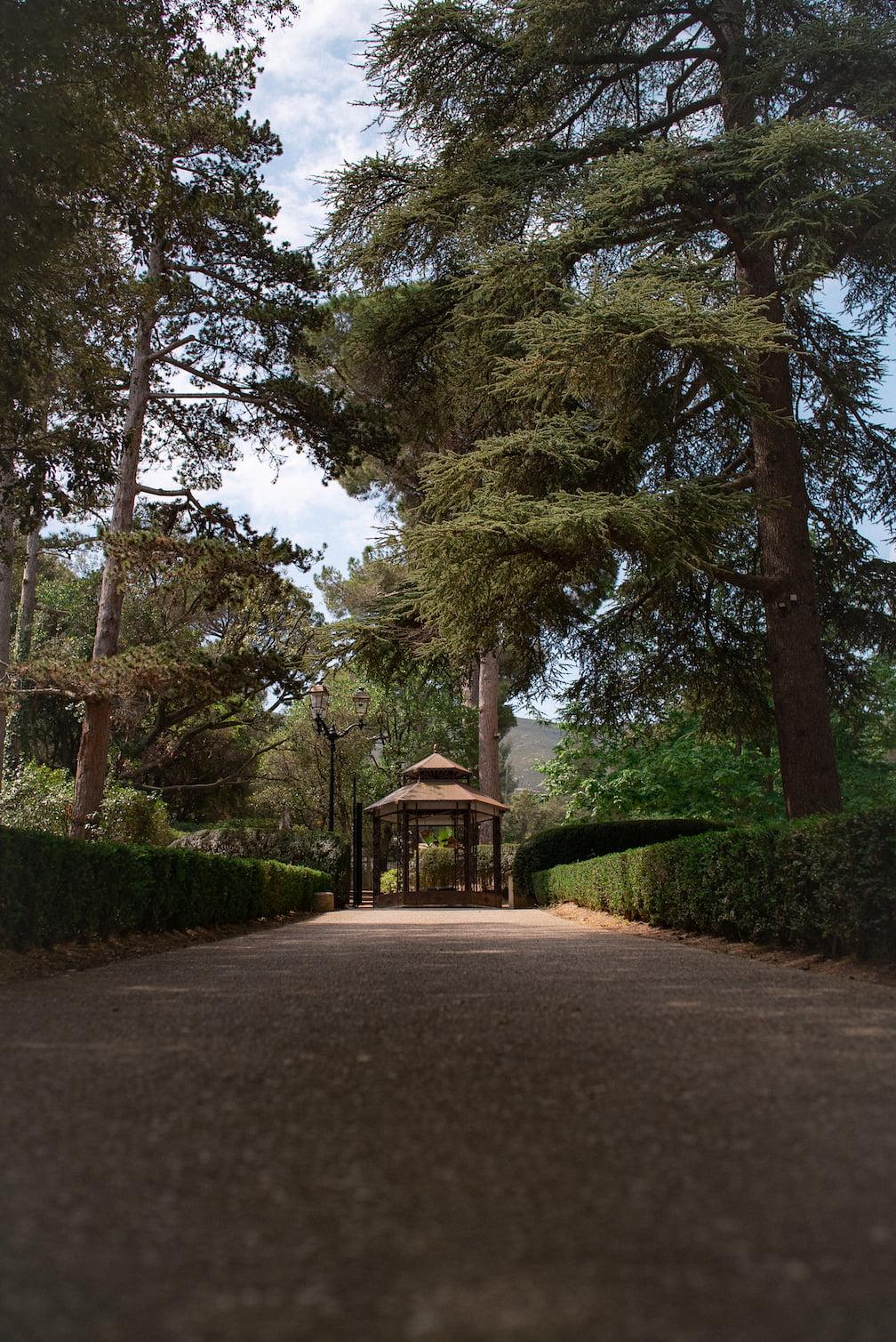 A serene path in a park leads to a picturesque gazebo surrounded by well-maintained hedges and tall lush trees. The sky is partly cloudy, creating a peaceful atmosphere.