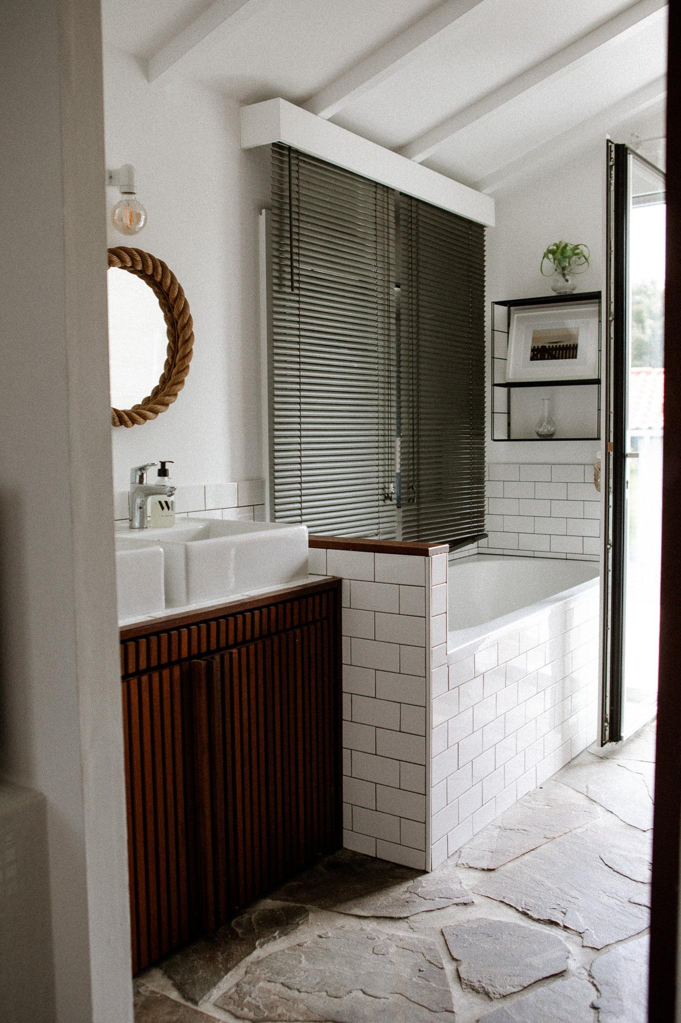 A bright bathroom with white tiles, wooden cabinets, a circular mirror with a rope frame, and a double Domino Uno sink. A bathtub sits next to black blinds covering a large window. The glass door is partially open, revealing shelves filled with various items, including a potted plant.