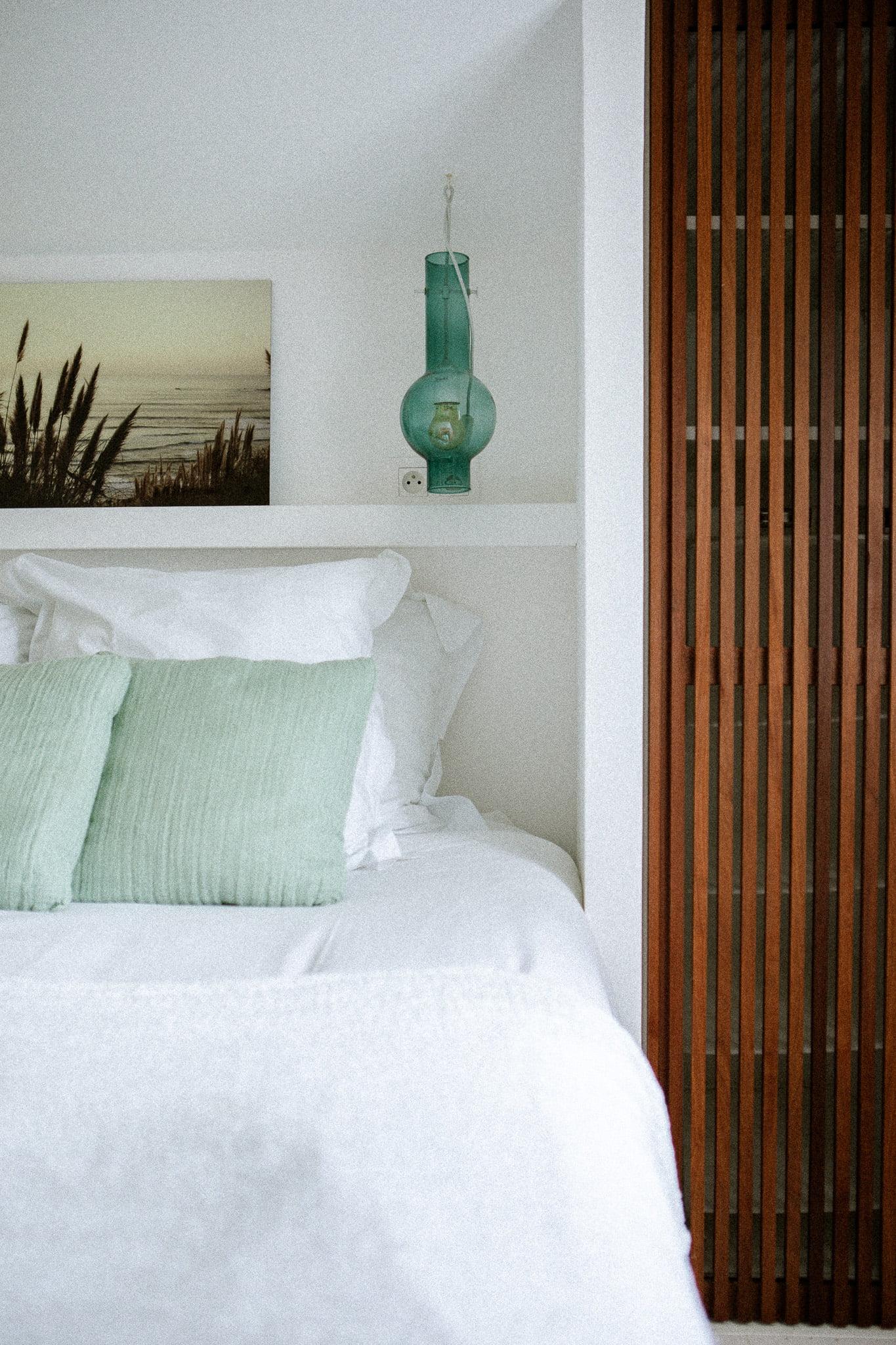 A minimalist bedroom features a white bed with two seafoam green pillows and white sheets. Above the bed, a shelf holds a turquoise glass vase and a framed coastal photograph. A wooden slatted closet door stands to the right, creating a serene and tidy atmosphere reminiscent of Domino Uno style.