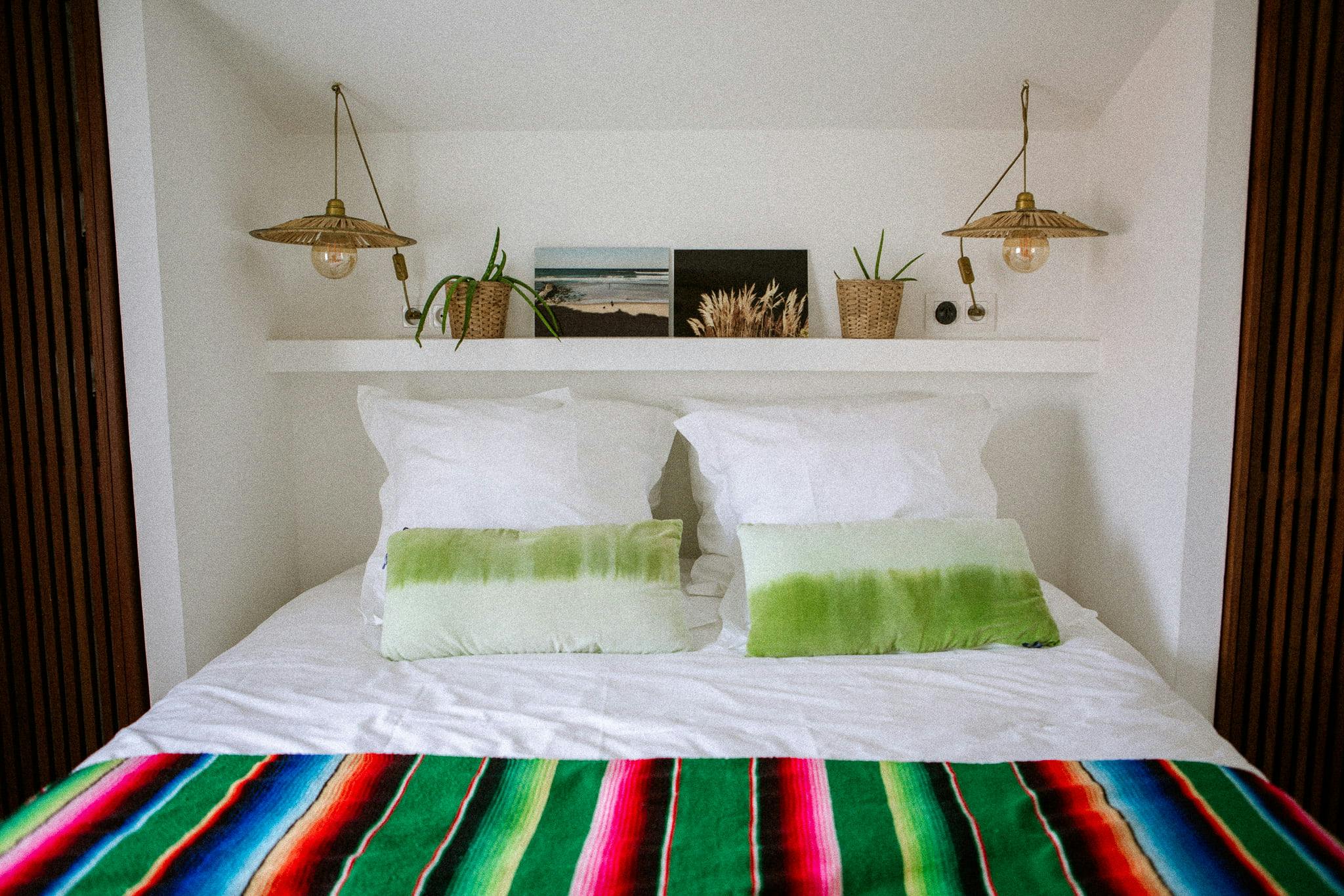 A cozy bedroom with a neatly made bed featuring white pillows and pillows with green accents. A vibrant striped Domino Uno blanket covers the lower half of the bed. The wall above the bed displays framed pictures, potted plants, and two hanging fixtures.