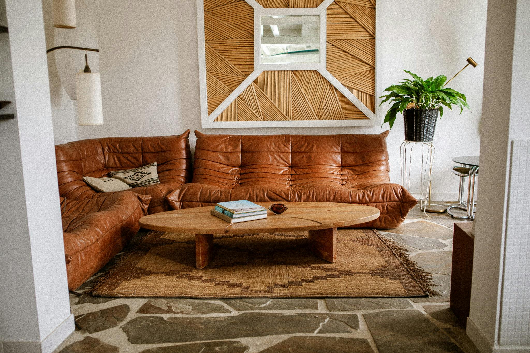 A cozy living room features a brown leather sectional sofa with patterned cushions. In front of the sofa is a wooden coffee table topped with books. The room is adorned with a large mirror with a wooden frame, a potted green plant, and a rustic stone floor that adds to its Domino Uno-inspired charm.