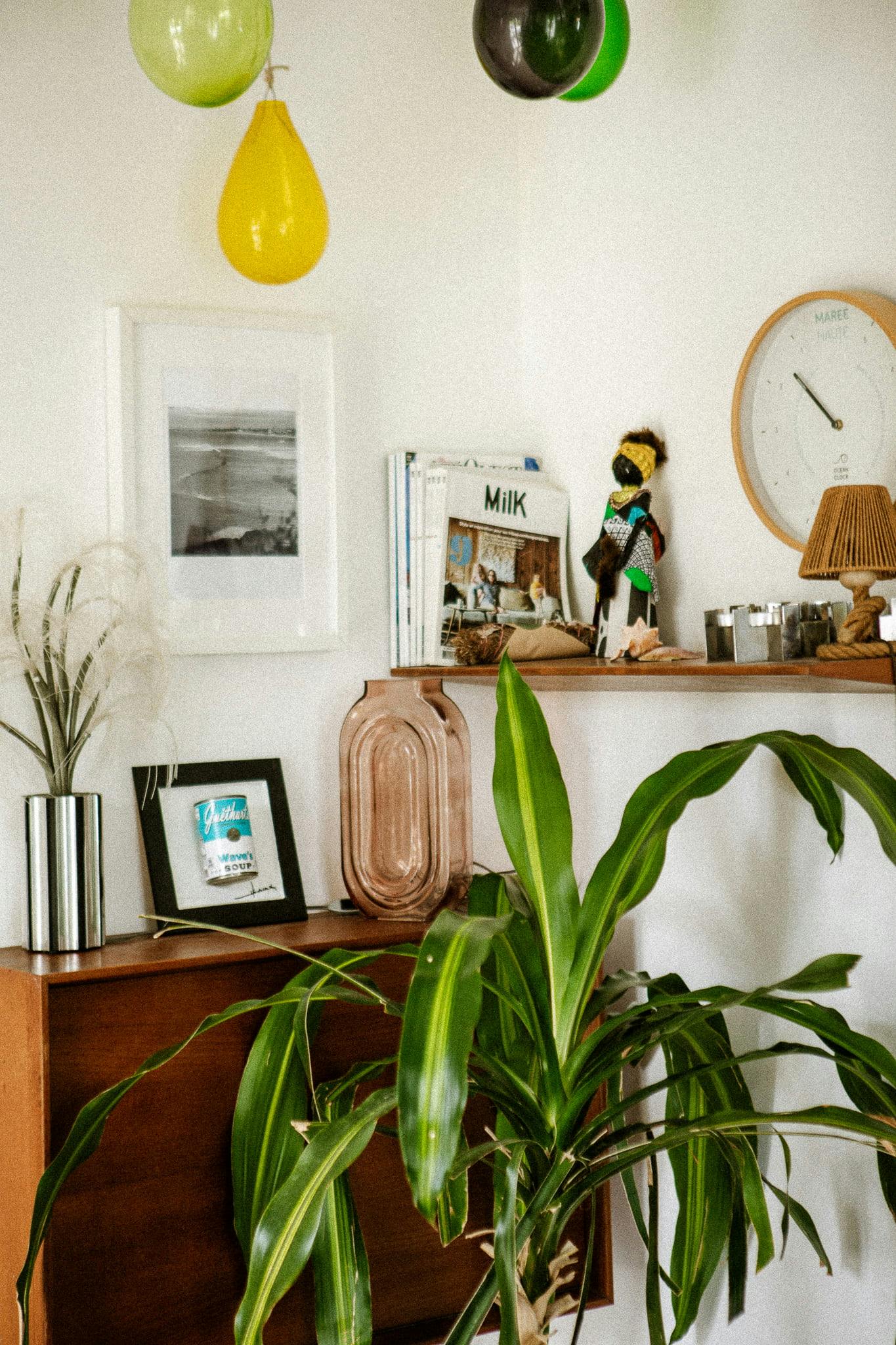 A cozy nook features leafy green plants, a small brown shelf filled with books and decorative items, and a round Domino Uno clock. Green and yellow balloons are suspended from the ceiling. A framed black-and-white photograph and a framed book cover are displayed on the wooden console beneath.