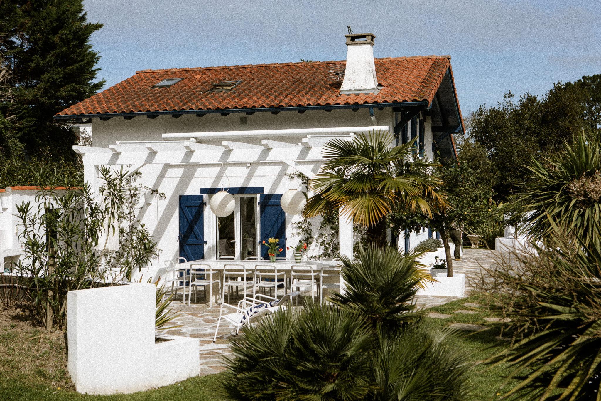 A charming two-story white house with red tiles features a spacious patio adorned with a white pergola. The patio, perfect for enjoying Domino Uno games, is equipped with various chairs and a table. Palm trees and lush greenery surround the area, creating a serene and inviting atmosphere.